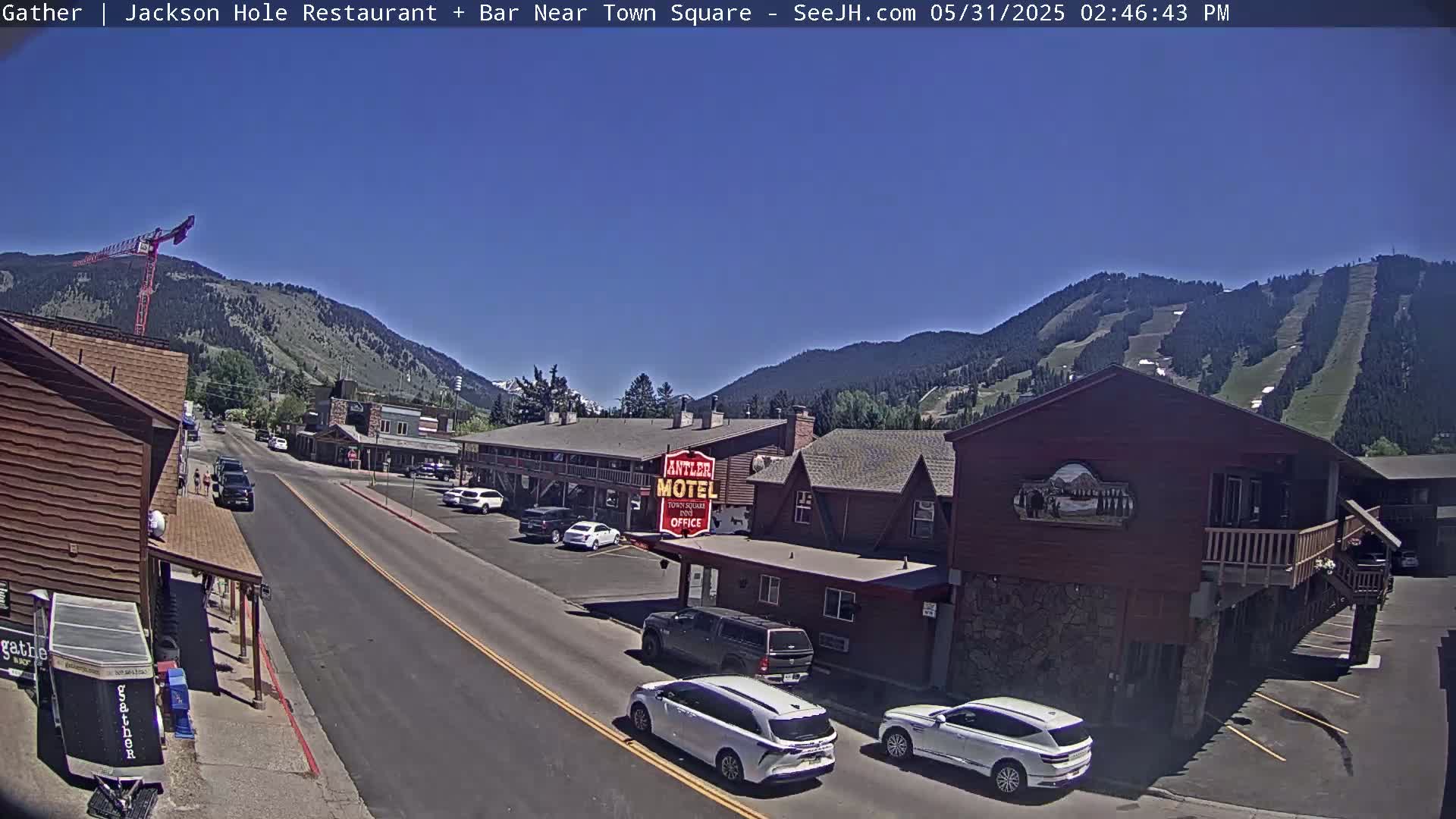 A sunny day reveals a town street scene nestled in a valley, with mountains in the background showing ski slopes and a construction crane.