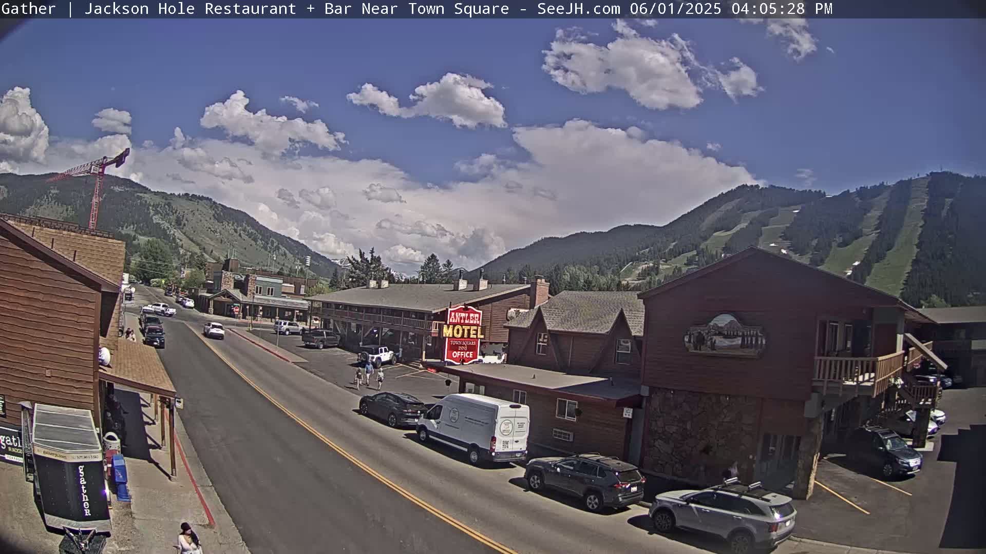 A partly sunny day shows a street scene in a mountain town with cars parked along the side of the road and buildings including a motel.