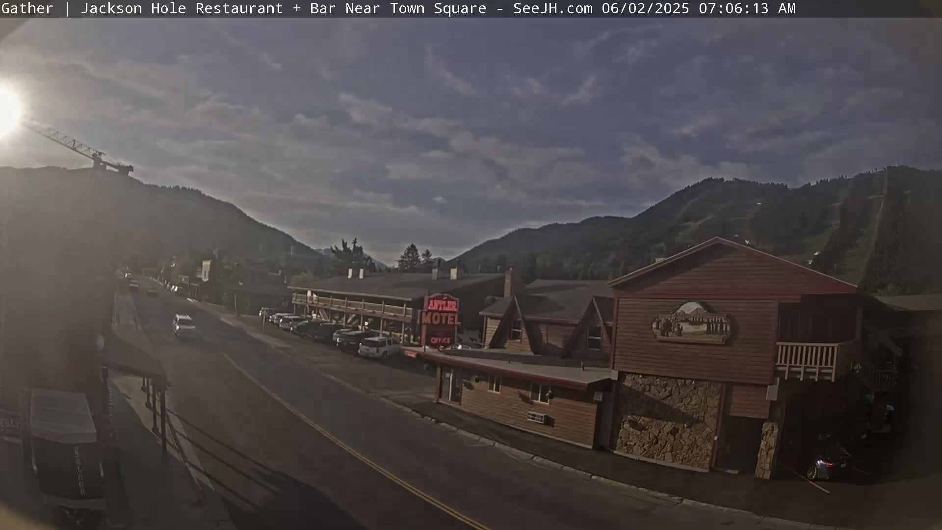 A partly cloudy morning scene shows a motel and street in a mountainous area, with cars parked along the roadside.