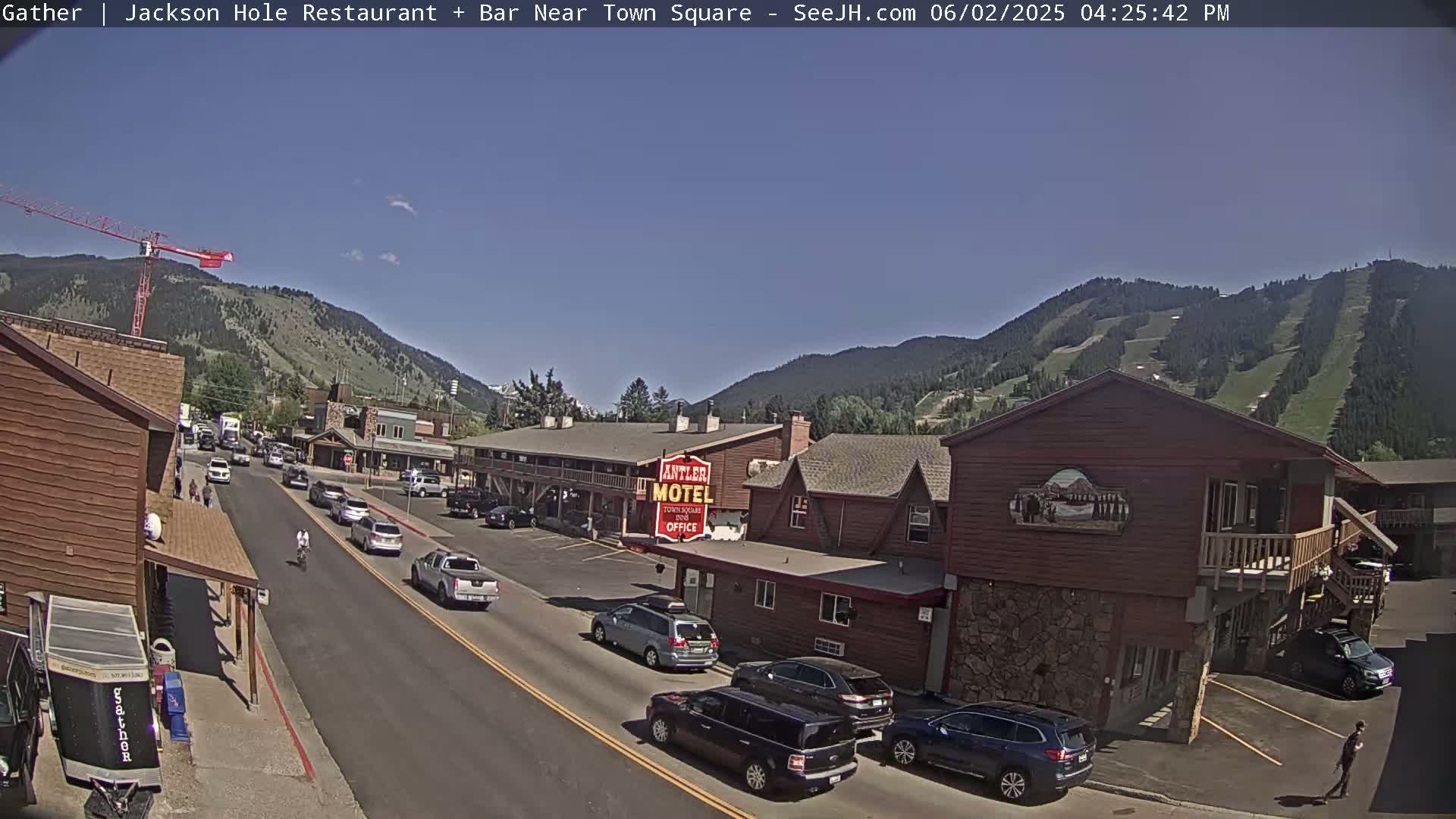 A sunny day reveals a street scene in a mountain town, with cars parked along the side of a road that leads toward a motel and a background of mountains partially covered in greenery.
