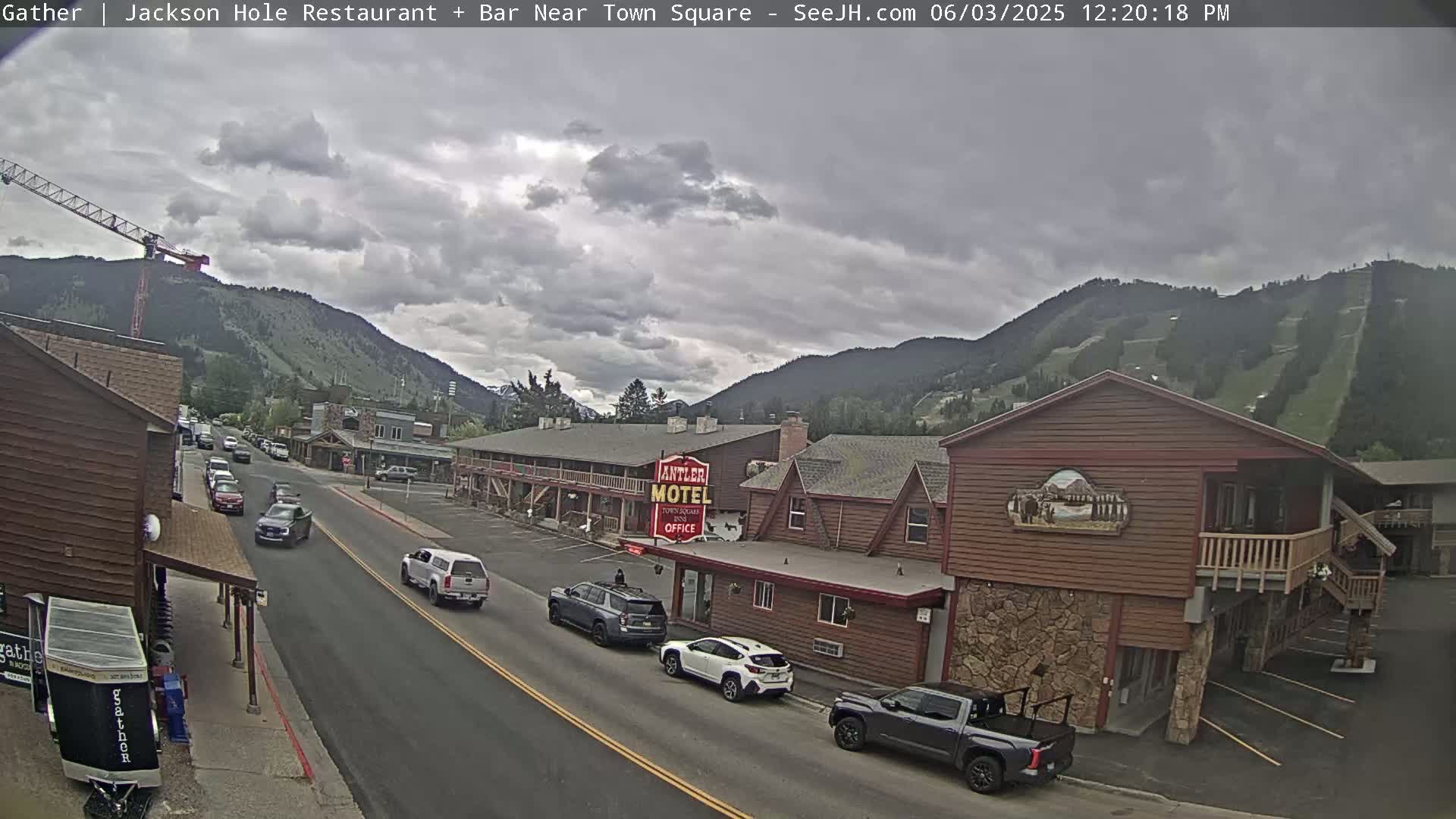 A partly cloudy day shows a street scene in a mountain town with cars driving past a motel and other buildings, and a construction crane visible in the distance.