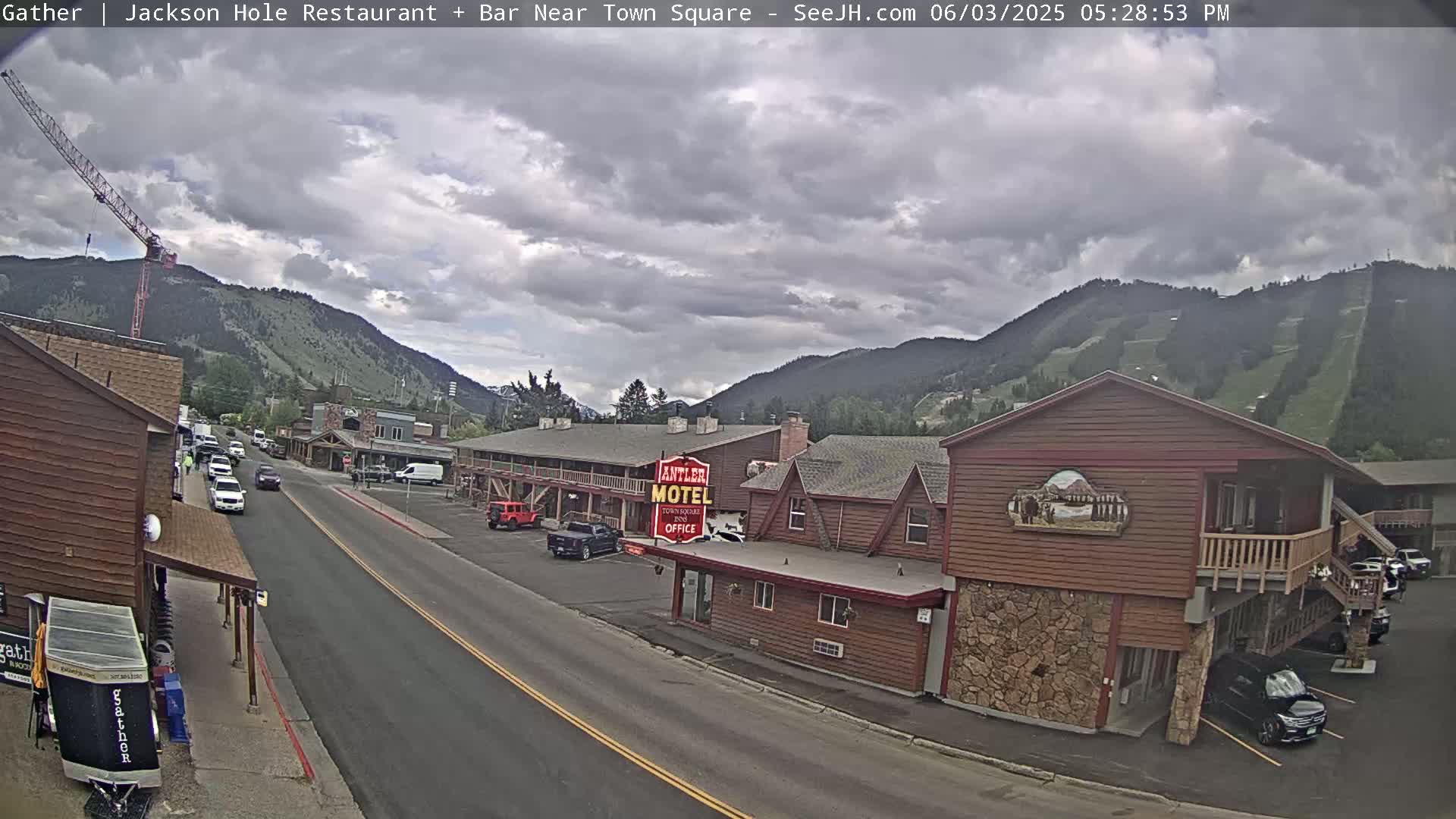 A street scene in a mountain town under a partly cloudy sky shows cars parked along a road in front of a motel and other buildings, with a construction crane visible in the distance.