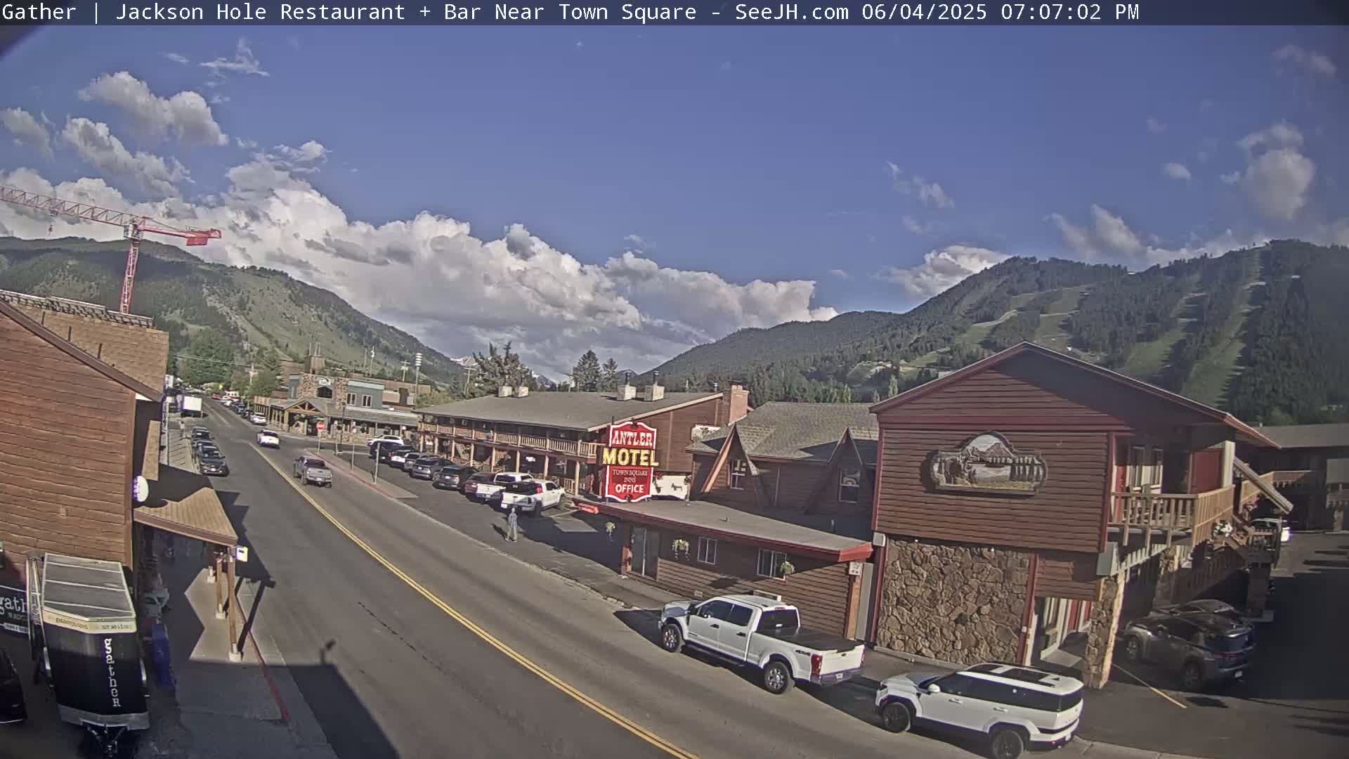 A partly sunny day reveals a street scene in a mountain town, with cars parked along the roadside in front of a motel and other buildings.