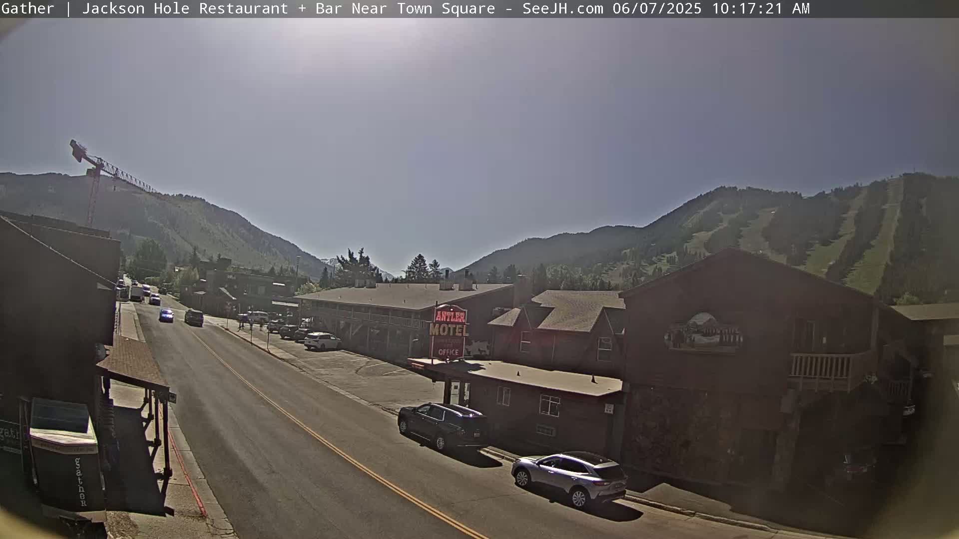A sunny day reveals a street scene in a mountain town, with cars parked along the roadside near a motel, and mountains visible in the background.