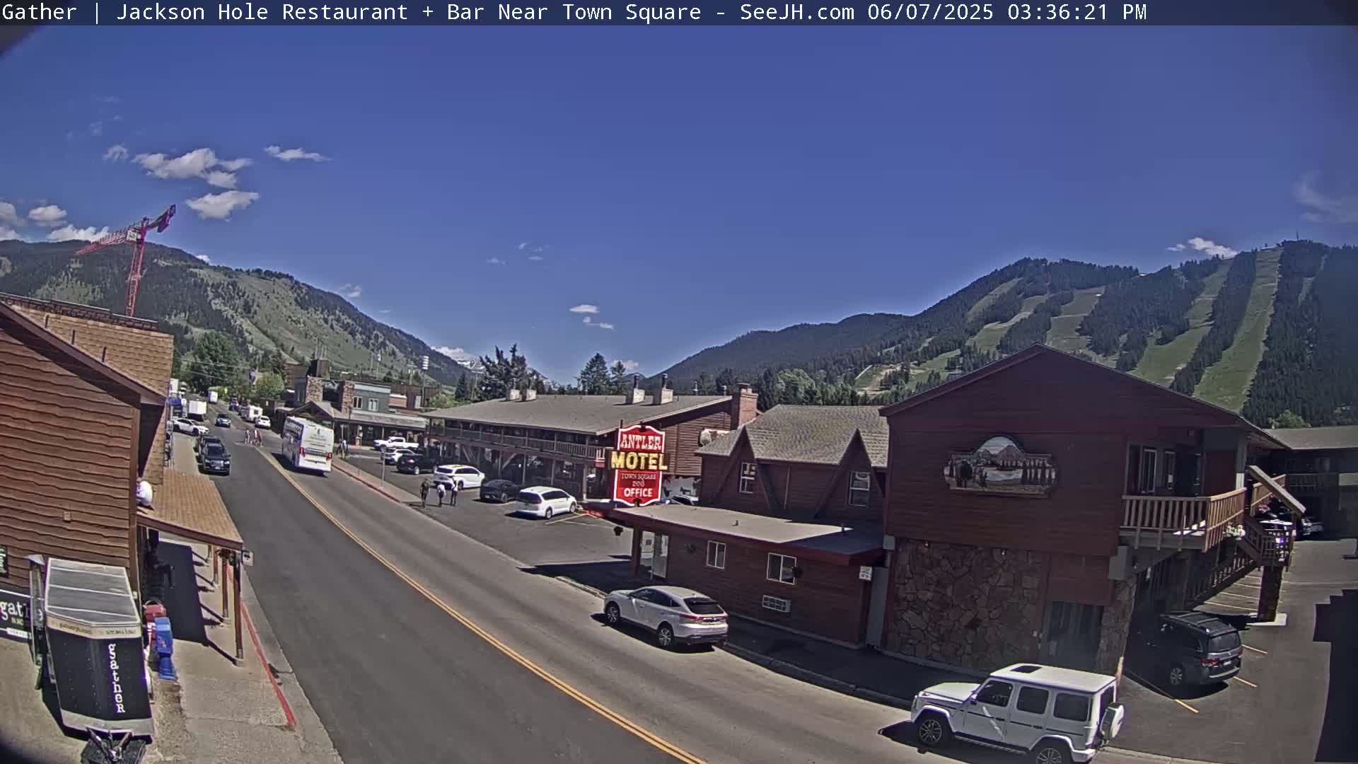 A sunny day reveals a street scene in a mountain town, with cars parked along the sides of a road lined with buildings, including a motel, and mountains in the background.