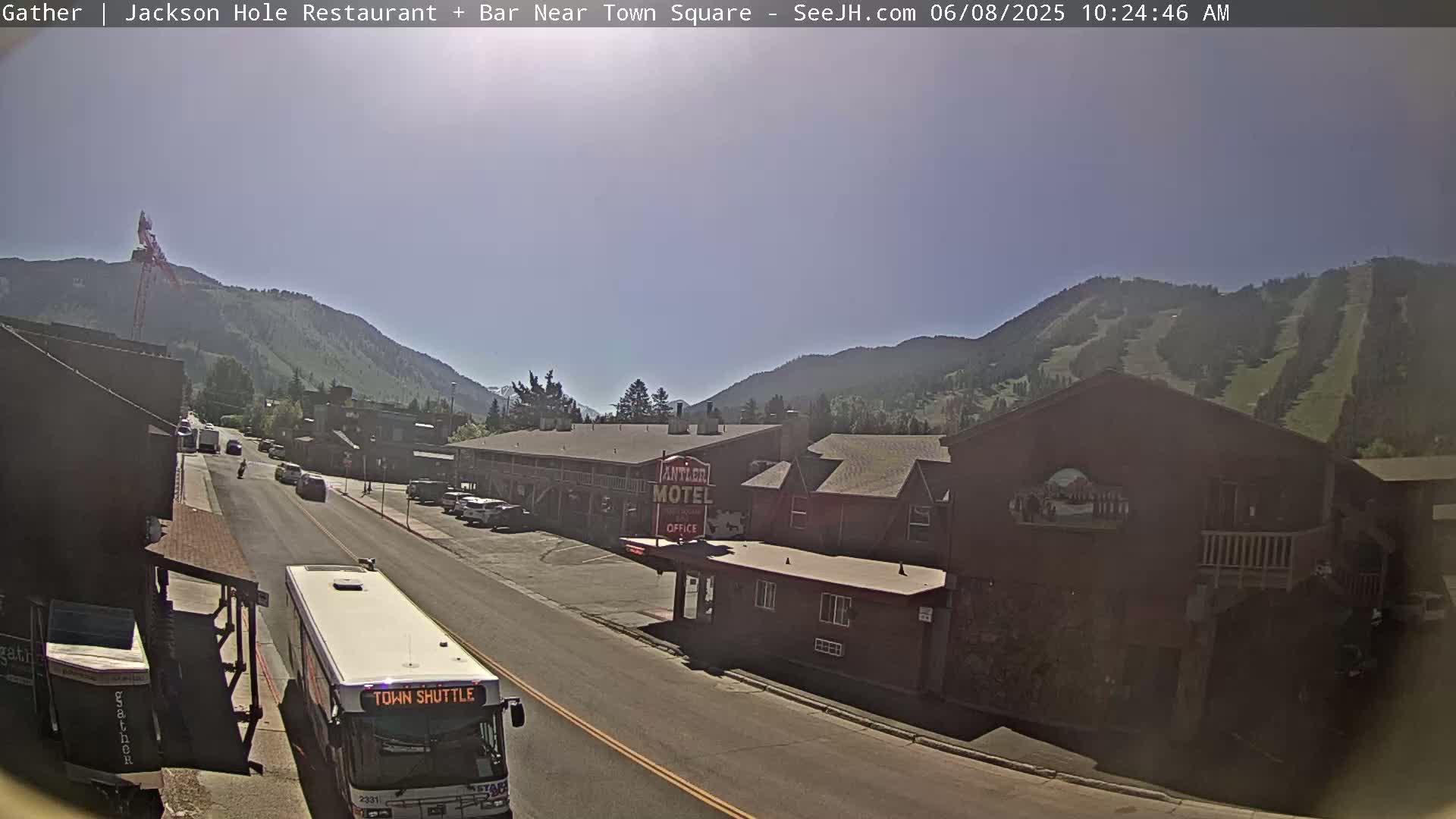 A town shuttle bus is parked on a street in front of a motel, with mountains in the background under a sunny sky.
