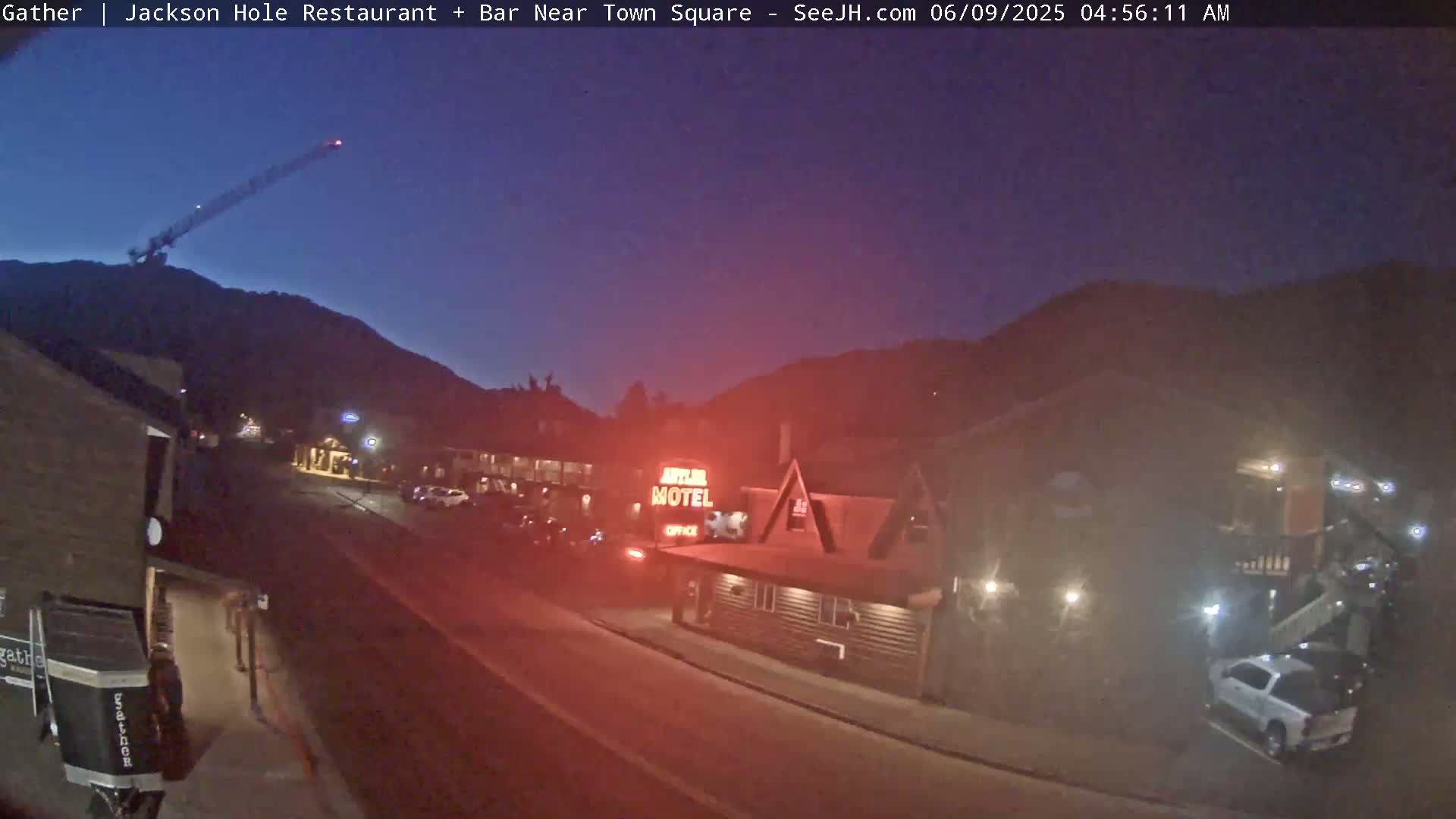 A nighttime street scene shows a motel and other buildings near a mountain under a twilight sky, with a crane visible in the distance.