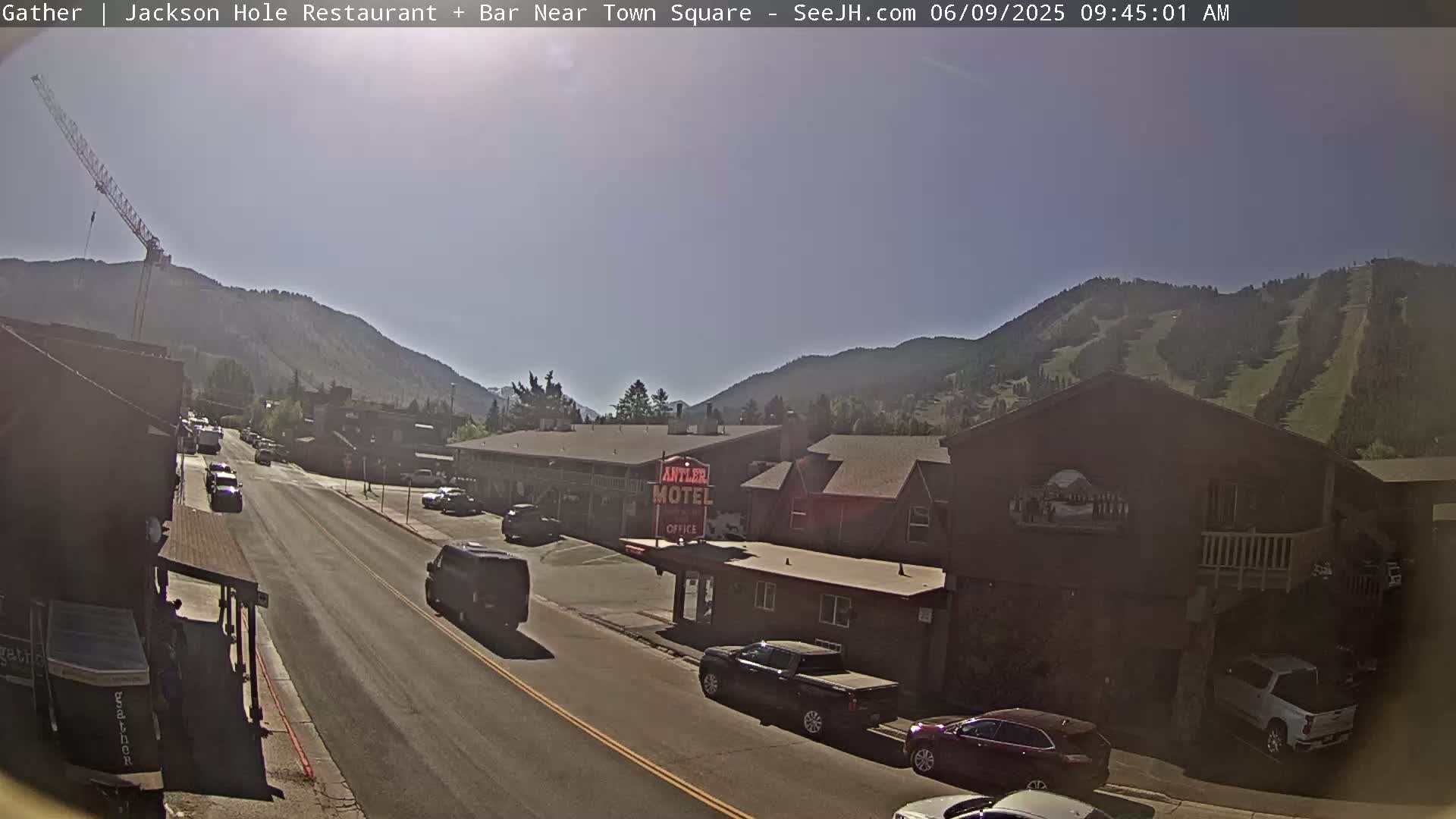 A sunny day reveals a street scene in a mountain town, with vehicles parked and driving past a motel, a construction crane visible in the distance against a backdrop of mountains.