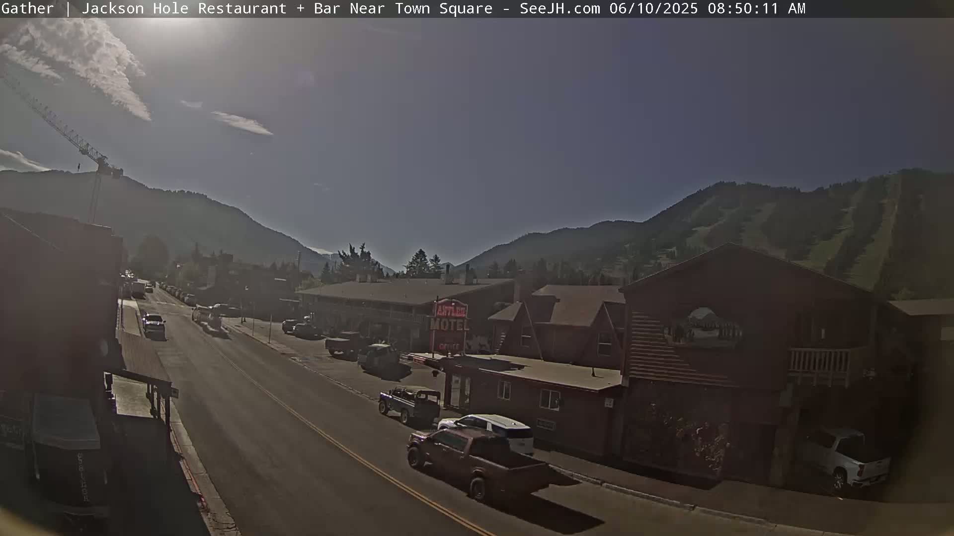 A sunny day reveals a street scene in a mountainous area with cars parked and driving along a road, and buildings including a motel in the foreground.