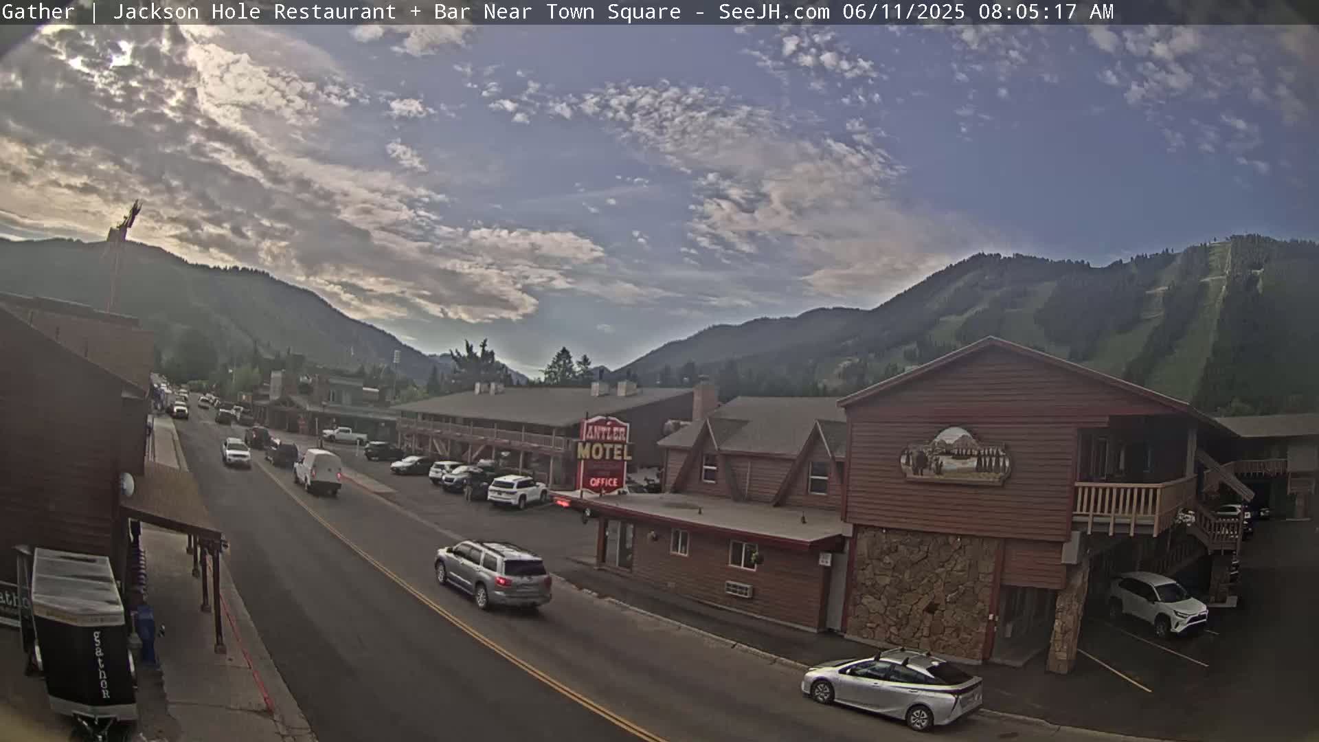 A partly cloudy morning scene shows a street in a mountain town with cars parked and driving, alongside a motel and other buildings, with mountains in the background.