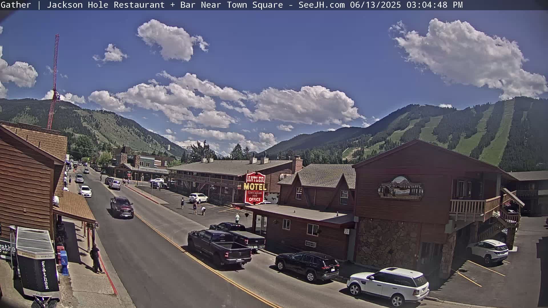 A partly sunny day shows a street scene in a mountain town with cars parked and driving, and buildings including a motel, under a partly cloudy sky with mountains in the background.