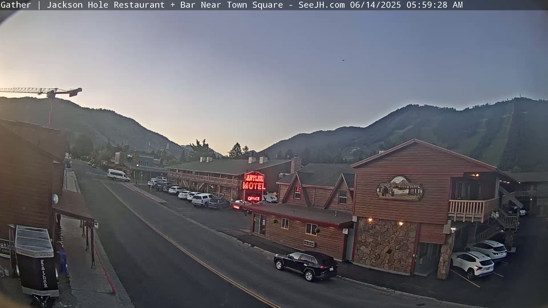A street scene at dawn shows a motel and other buildings near a mountain range under a clear, light-grey sky.