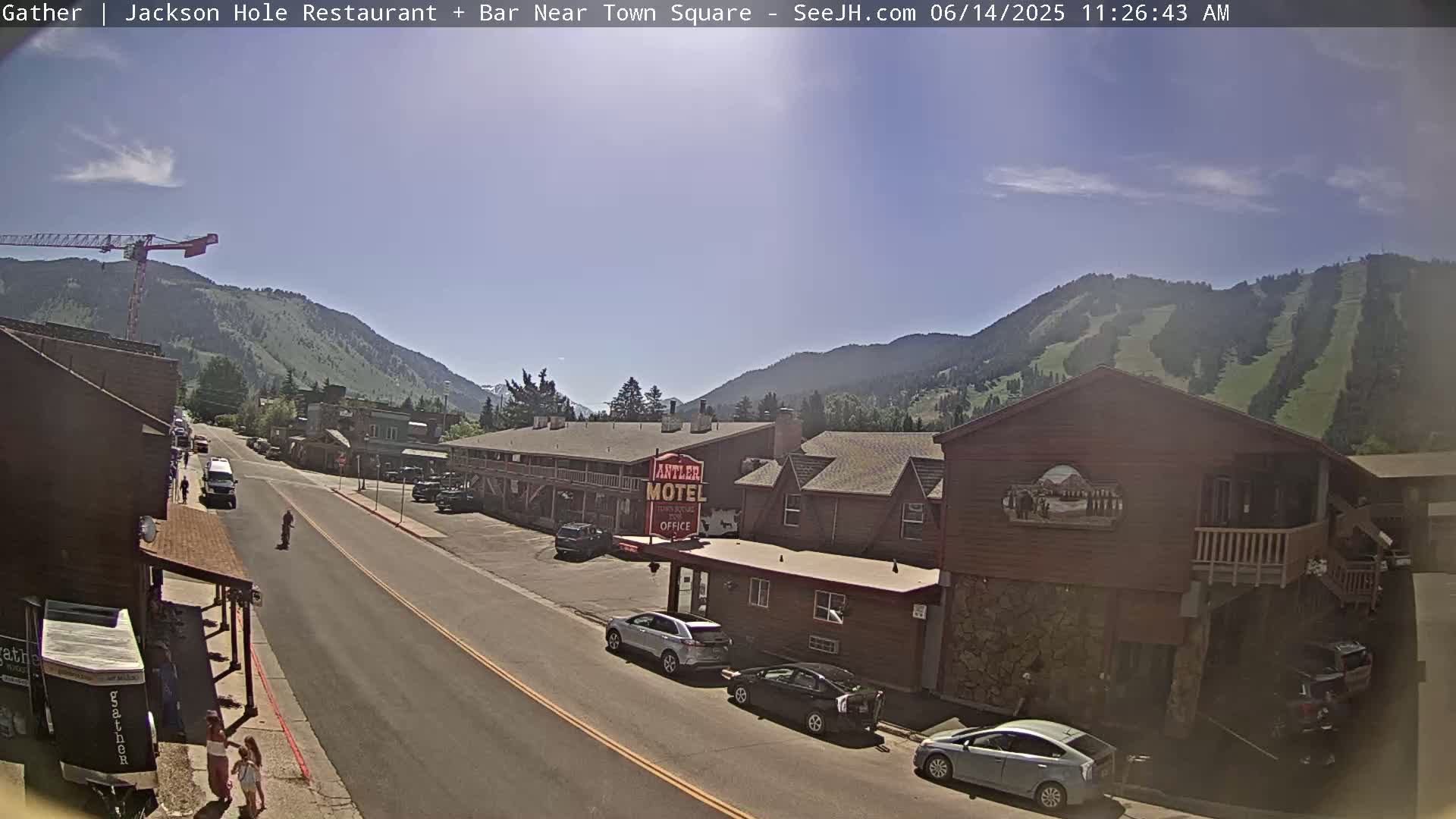 A sunny day reveals a street scene in a mountain town, with cars parked along the sides of buildings, a motel visible, and a construction crane in the distance.