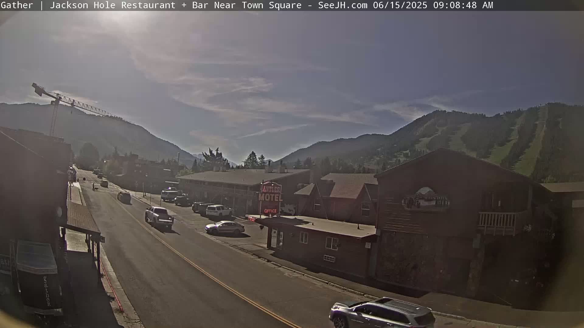A sunny day reveals a street scene with cars parked and driving along a road lined with buildings, including a motel, nestled against a mountain backdrop.