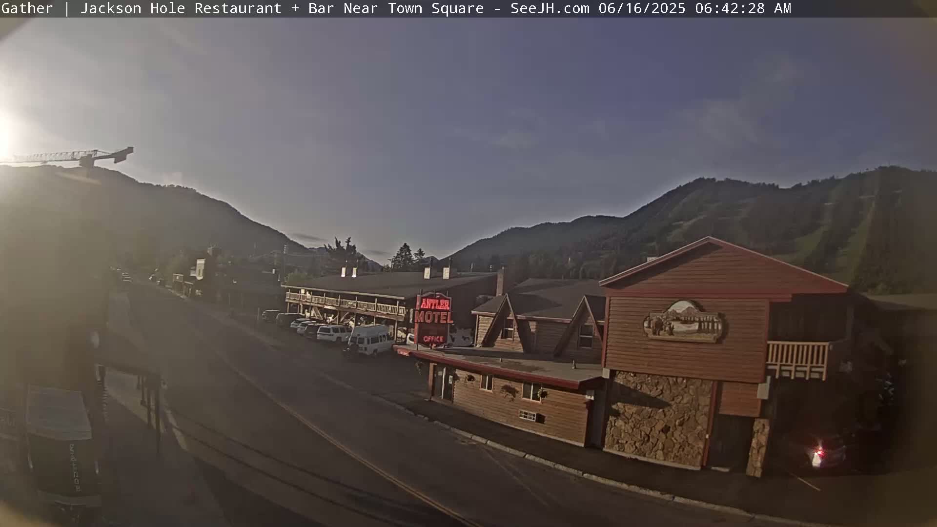 A sunlit street scene shows a motel and other buildings nestled at the base of a mountain range under a clear, mostly sunny sky.