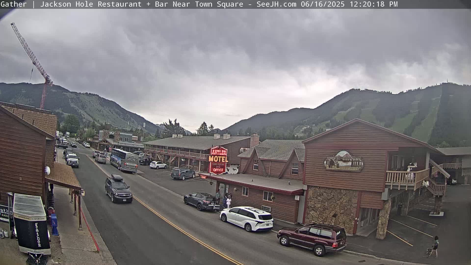 A street scene under an overcast sky shows a two-lane road with cars parked and driving past a motel and other buildings, with mountains in the background and a construction crane visible on the left.
