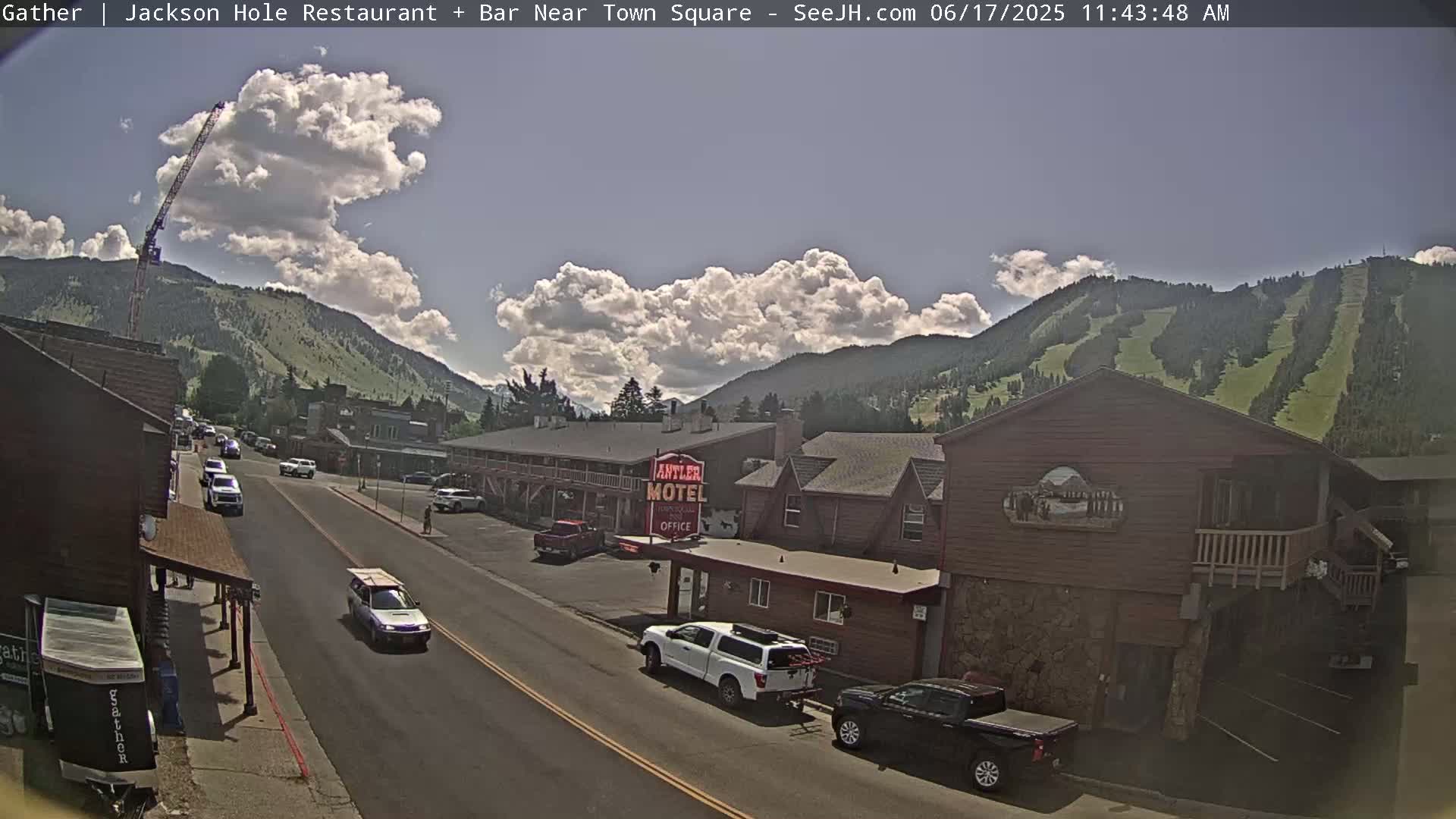 A partly cloudy day reveals a town street scene nestled in a valley, with mountains and ski slopes visible in the background; a construction crane is seen to the left.