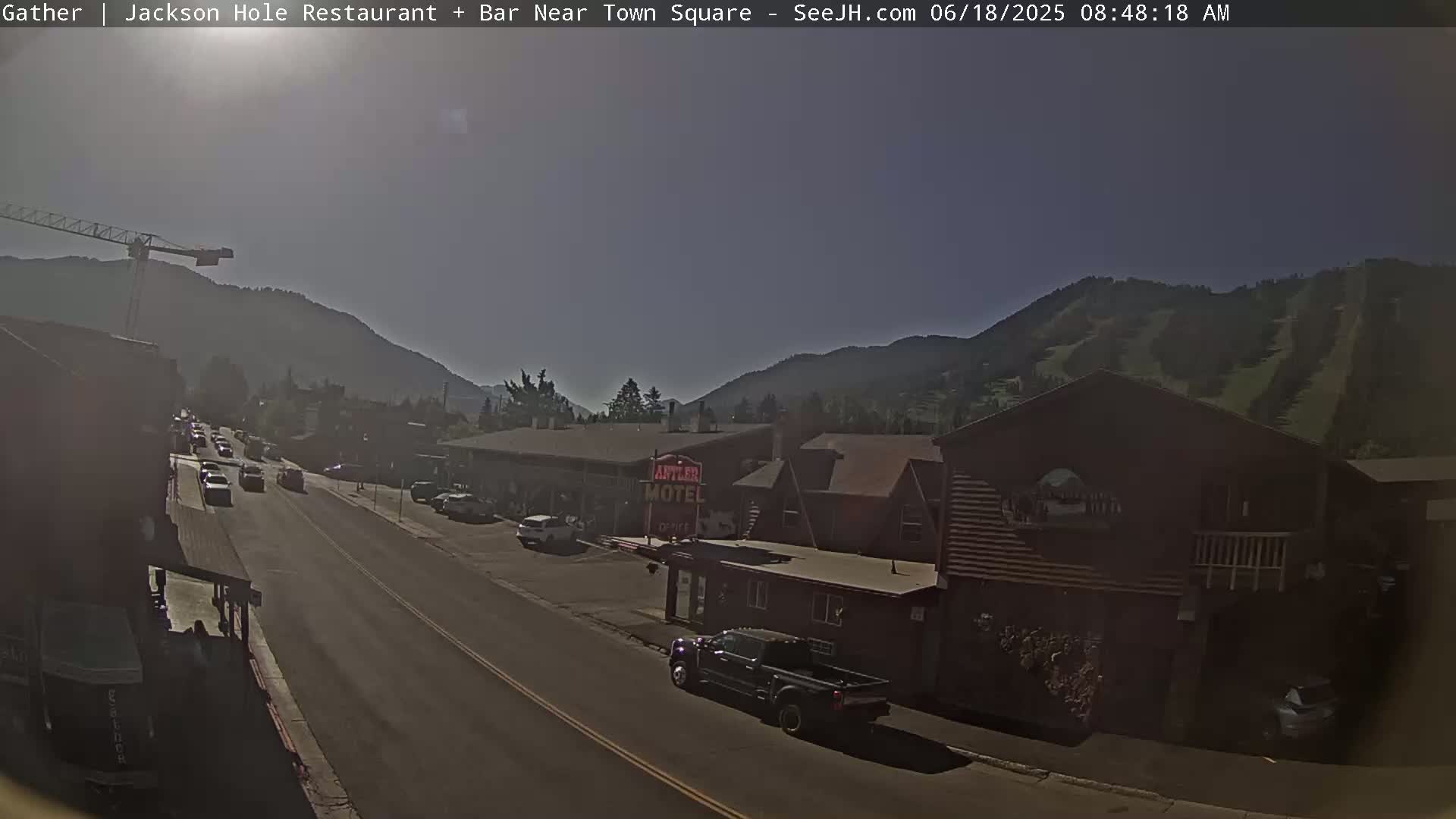 A sunny day reveals a street scene in a town nestled in a valley between mountains, with cars parked along the roadside and a motel visible.