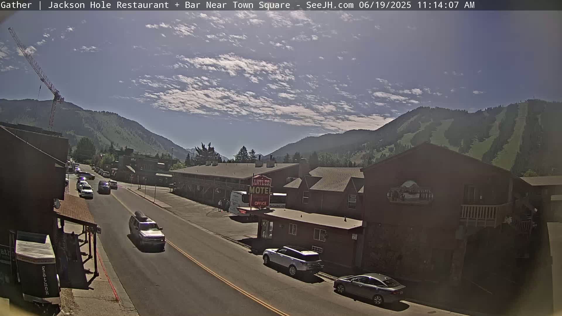A partly cloudy day shows a street scene in a mountain town with cars driving on a road, buildings, and a motel, along with a construction crane in the distance.