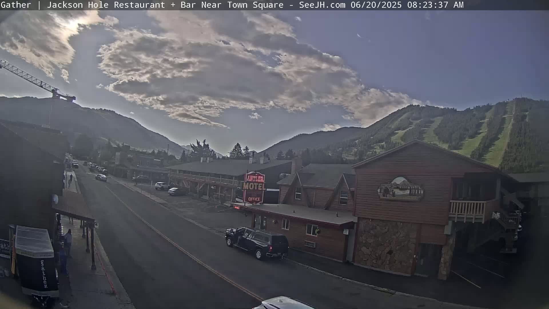 A partly cloudy morning scene shows a street with cars parked along the sides, buildings including a motel, and a mountain with visible ski slopes in the background.