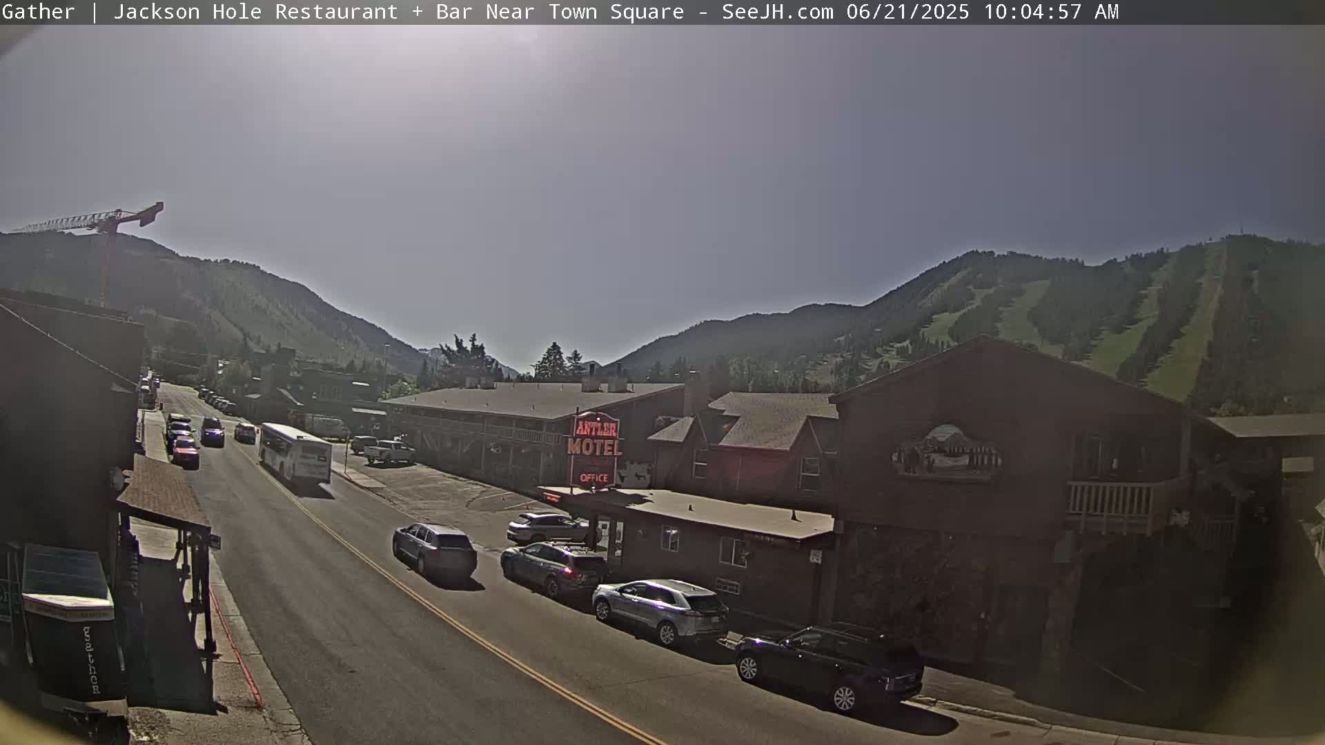 A sunny day reveals a street scene with cars and a bus traveling along a road, lined with buildings including a motel, and a mountainside with visible ski runs in the background.