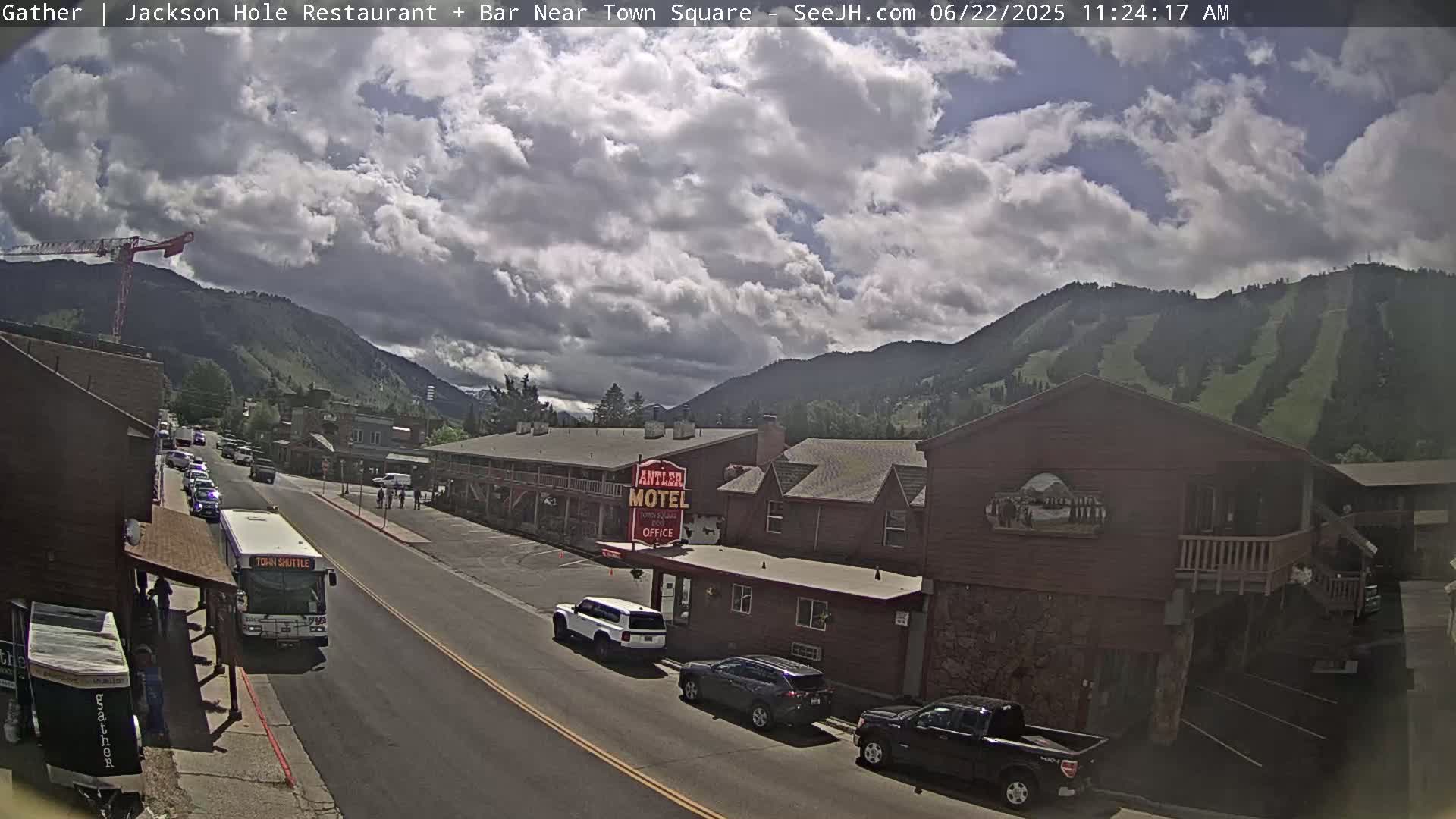 A partly cloudy day reveals a street scene in a mountainous town, with cars parked along the roadside near a motel and a bus at a bus stop.