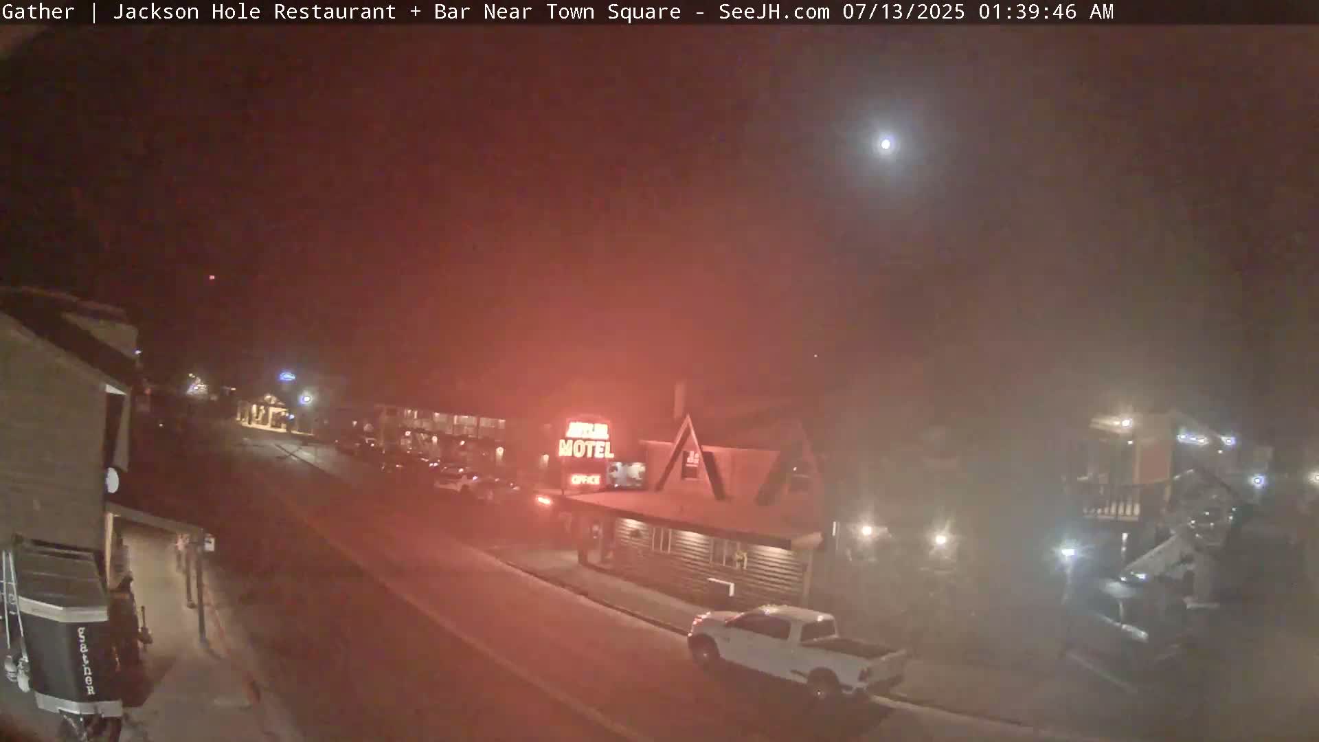 A foggy night scene shows a motel and a few parked cars along a street, with a bright light visible in the sky.