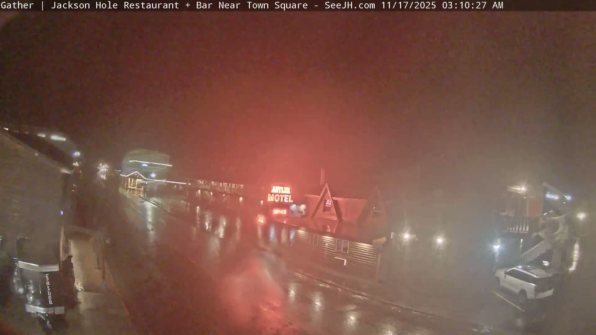 The image depicts a very dark and extremely foggy or misty night scene of a town street, wet from rain, featuring glowing building lights, the "Antler Motel" sign, and a few visible vehicles.