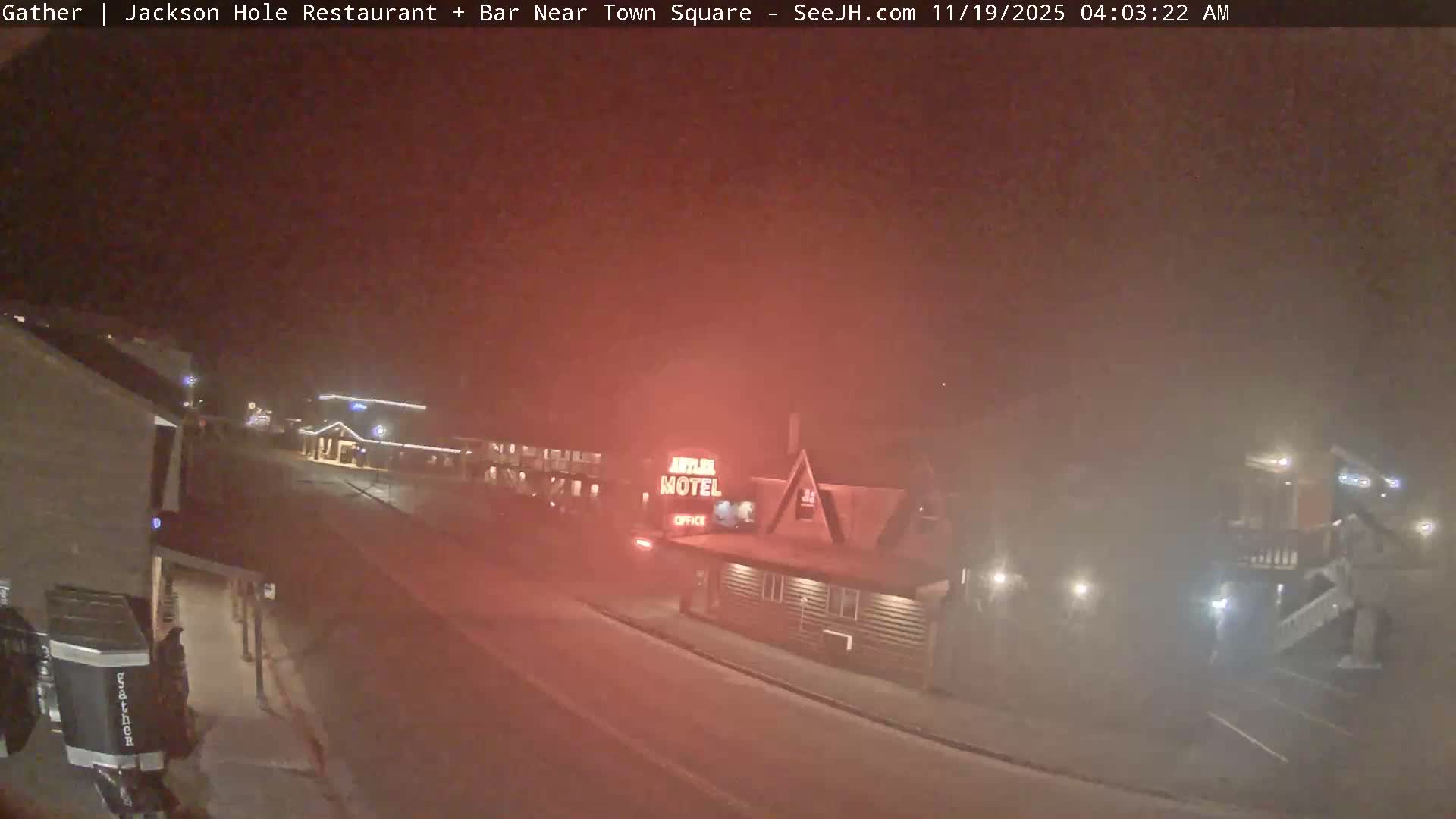 The image depicts a very dark and extremely foggy or misty night scene of a town street, wet from rain, featuring glowing building lights, the "Antler Motel" sign, and a few visible vehicles.