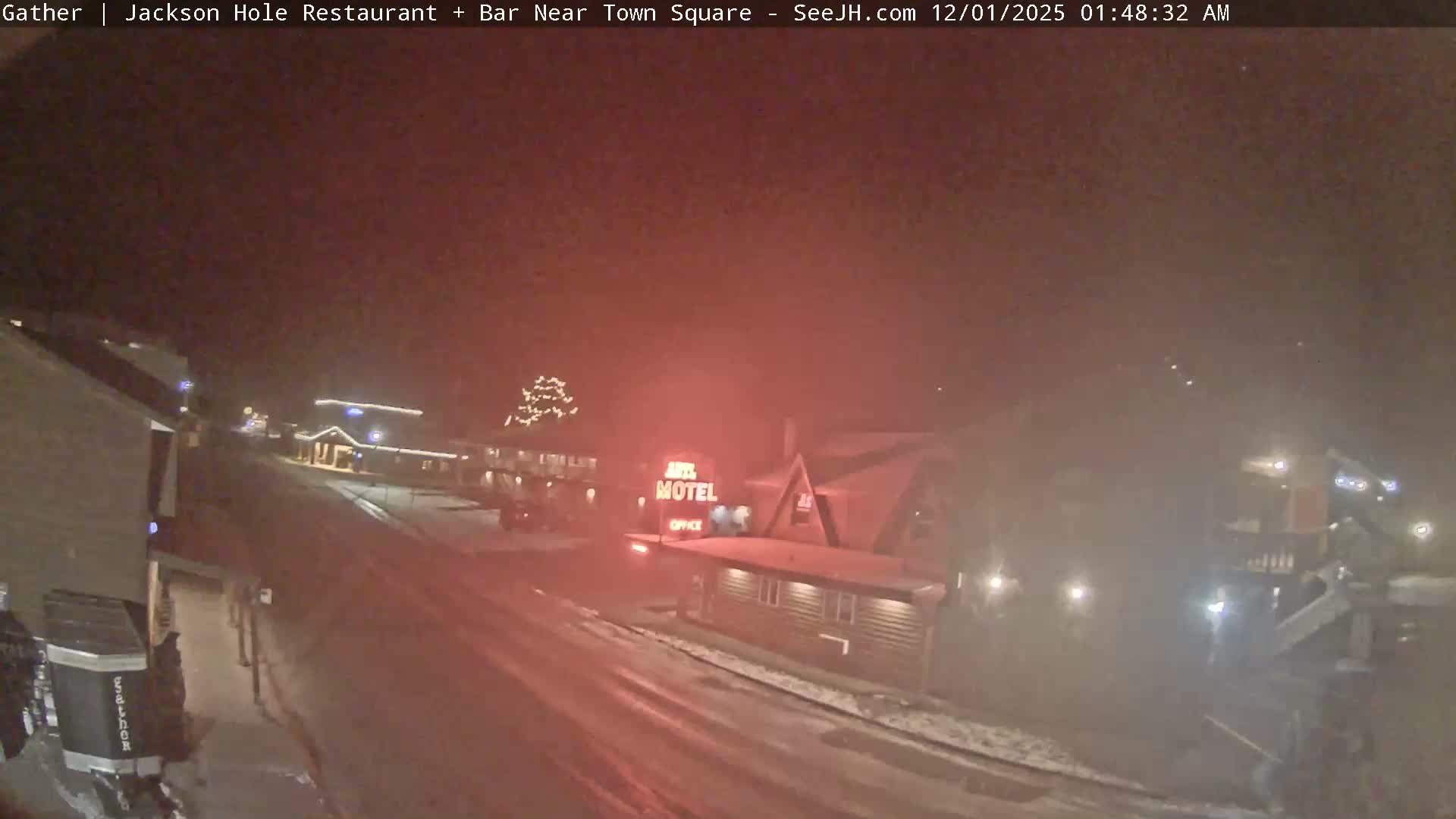 A hazy and blurry outdoor image taken at night reveals a snowy winter scene with falling precipitation, featuring a street, snow-dusted buildings decorated with festive lights, and a prominent red glowing sign.