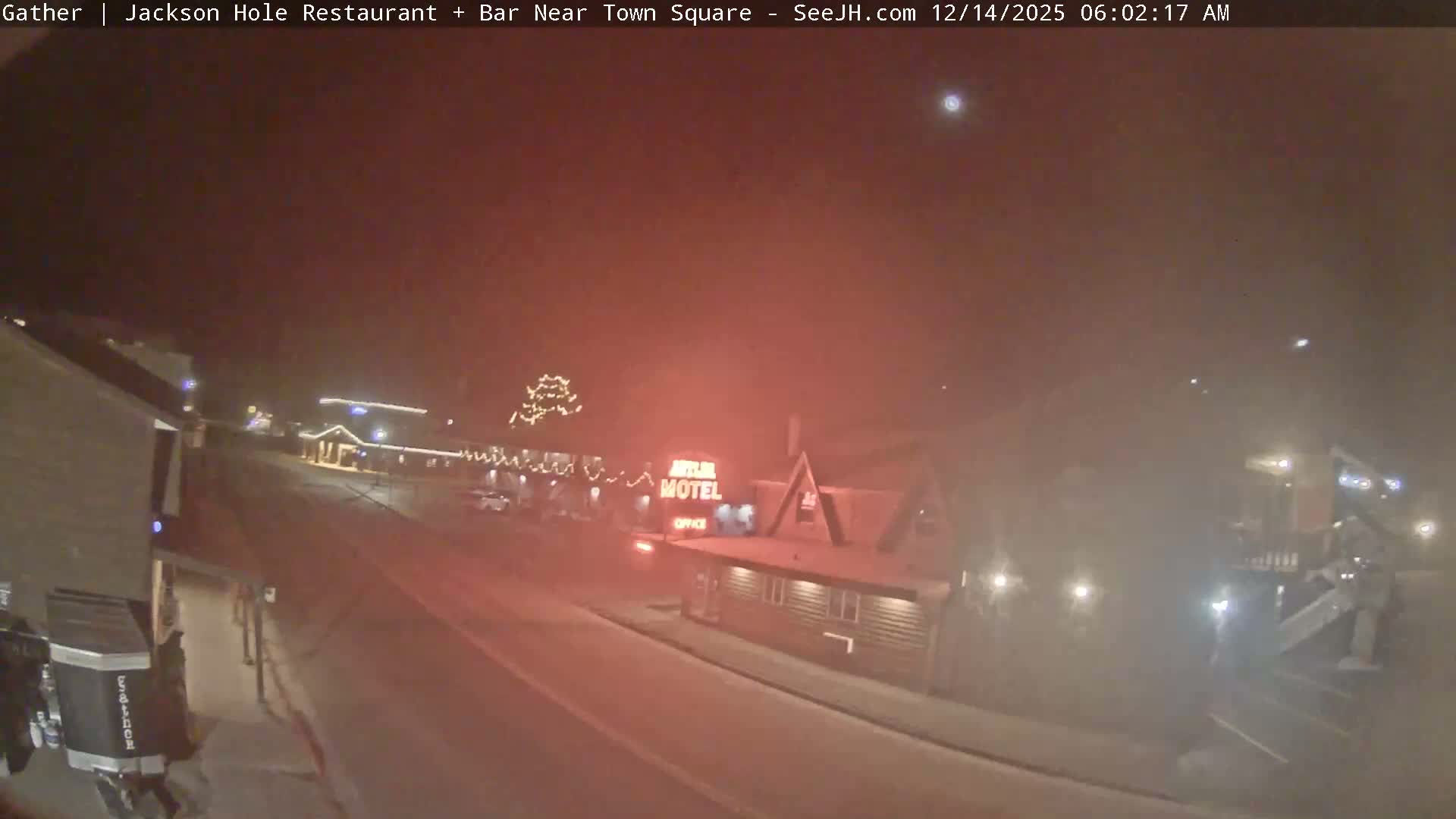 A hazy and blurry outdoor image taken at night reveals a snowy winter scene with falling precipitation, featuring a street, snow-dusted buildings decorated with festive lights, and a prominent red glowing sign.