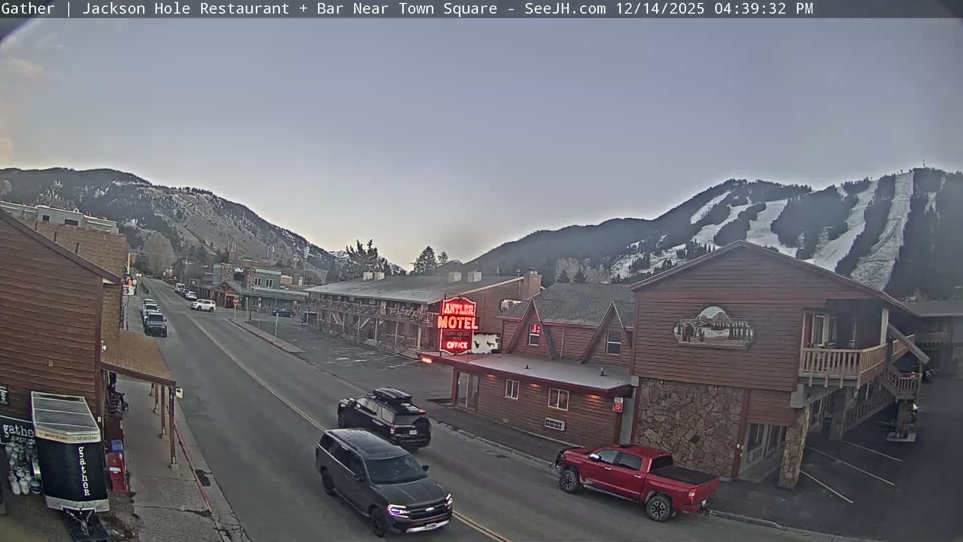 A hazy and blurry outdoor image taken at night reveals a snowy winter scene with falling precipitation, featuring a street, snow-dusted buildings decorated with festive lights, and a prominent red glowing sign.