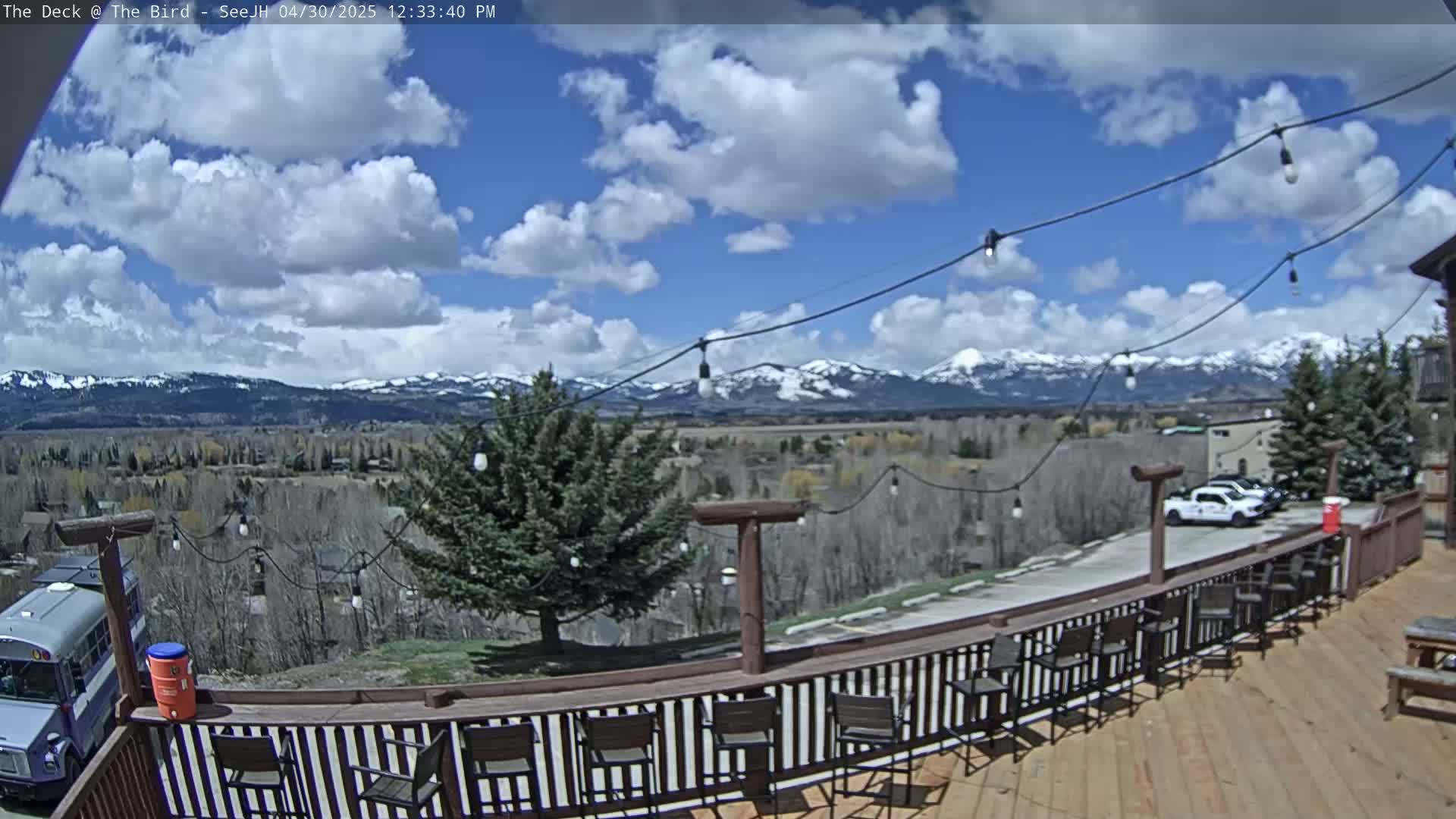 A partly cloudy day reveals snow-capped mountains in the distance, viewed from a wooden deck with chairs and string lights.