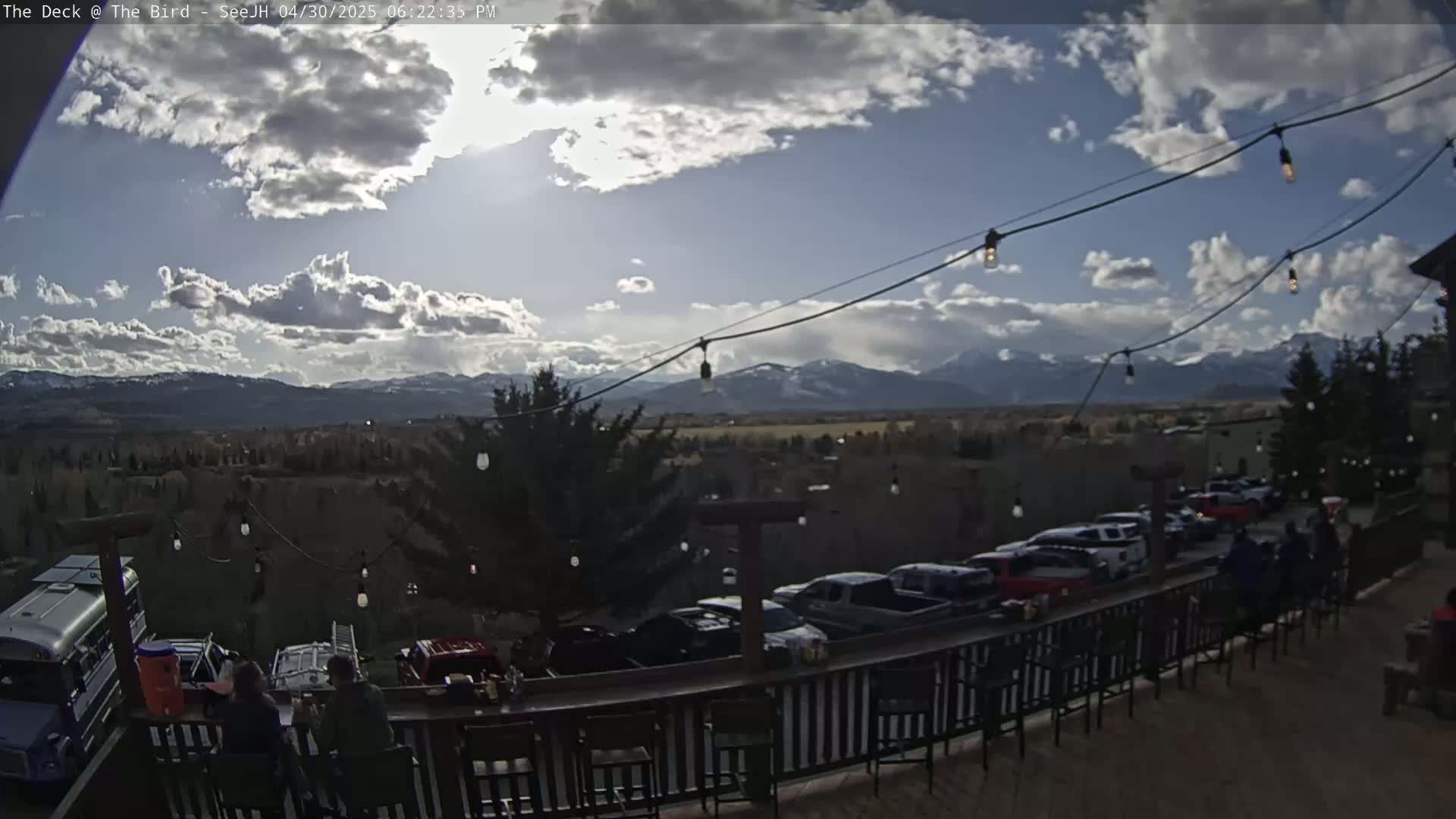 A partly cloudy outdoor scene shows a deck with people sitting at a bar overlooking a parking lot, a valley, and mountains in the distance.