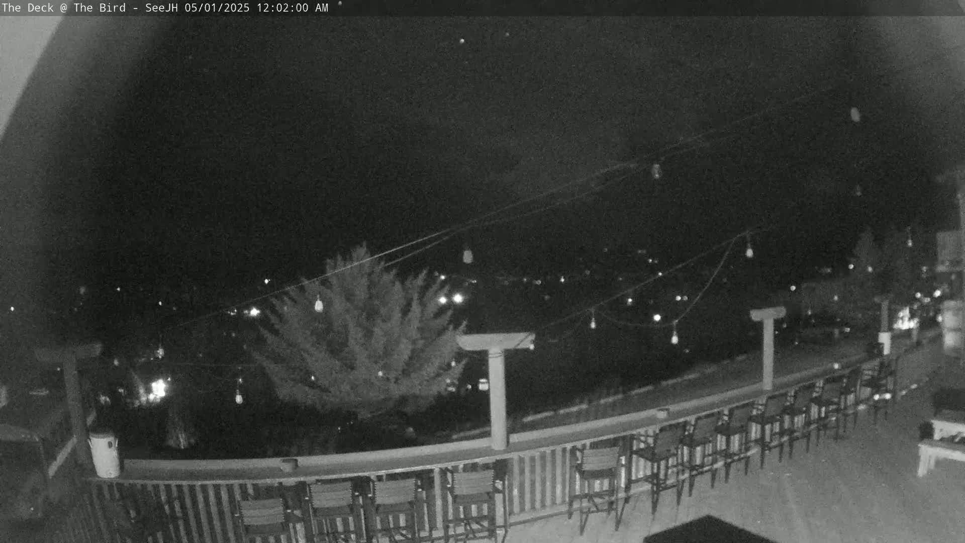 A nighttime view from a deck shows a row of empty chairs along a railing overlooking a town with string lights overhead.
