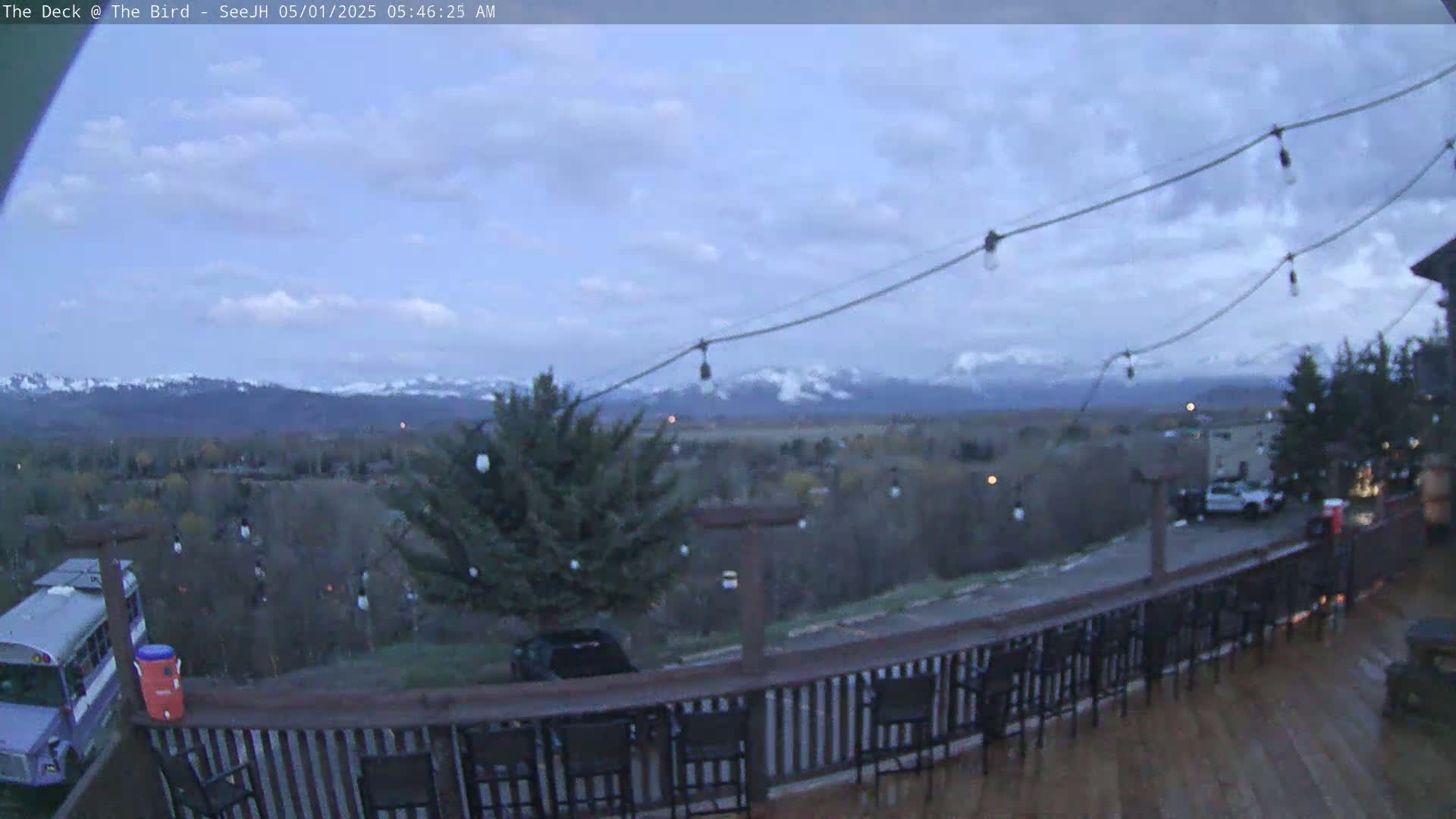 A deck with string lights overlooks a valley and snow-capped mountains under a cloudy sky.