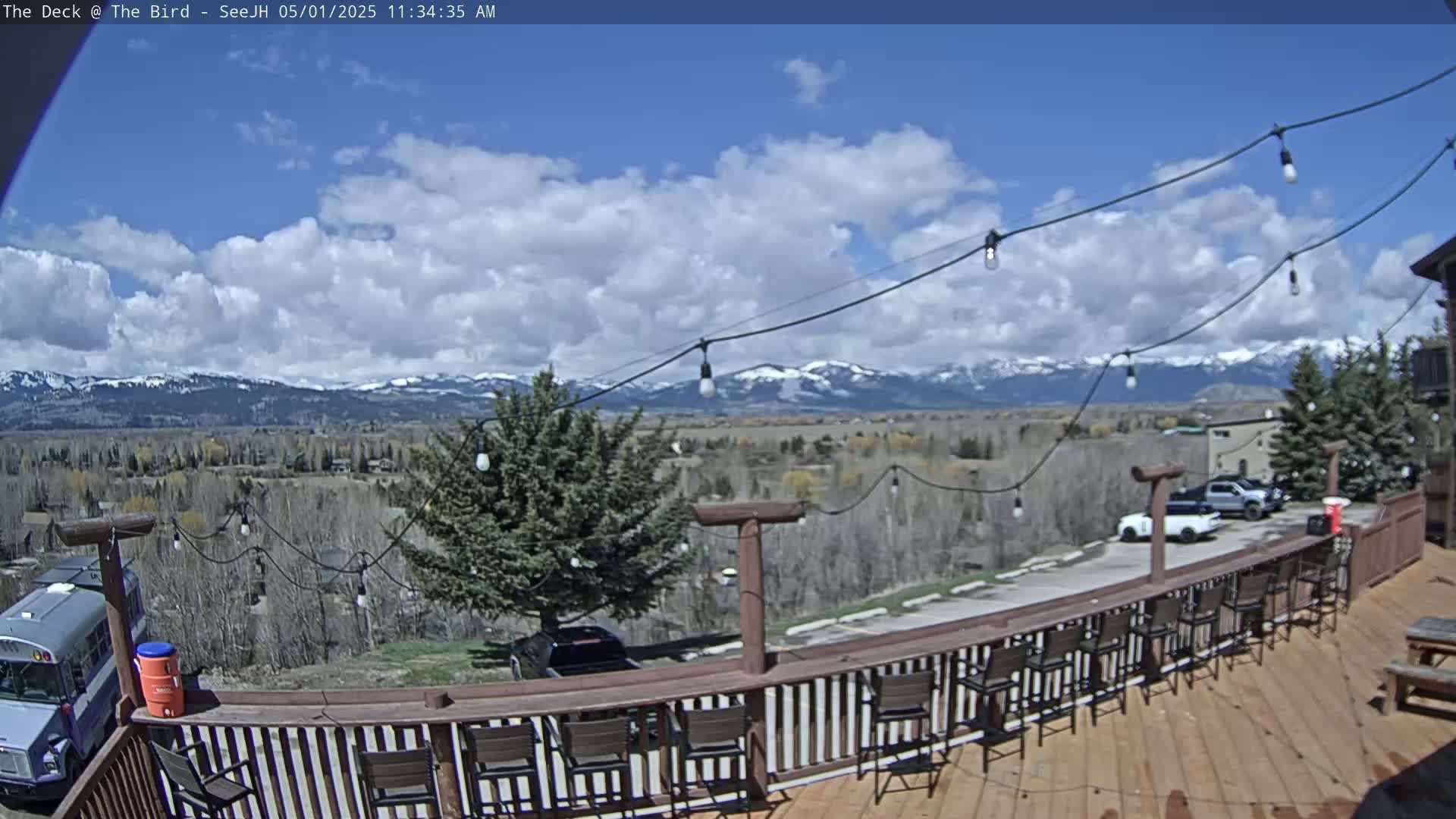 A deck with chairs and string lights overlooks a valley, a parking lot with vehicles, and snow-capped mountains under a partly cloudy sky.