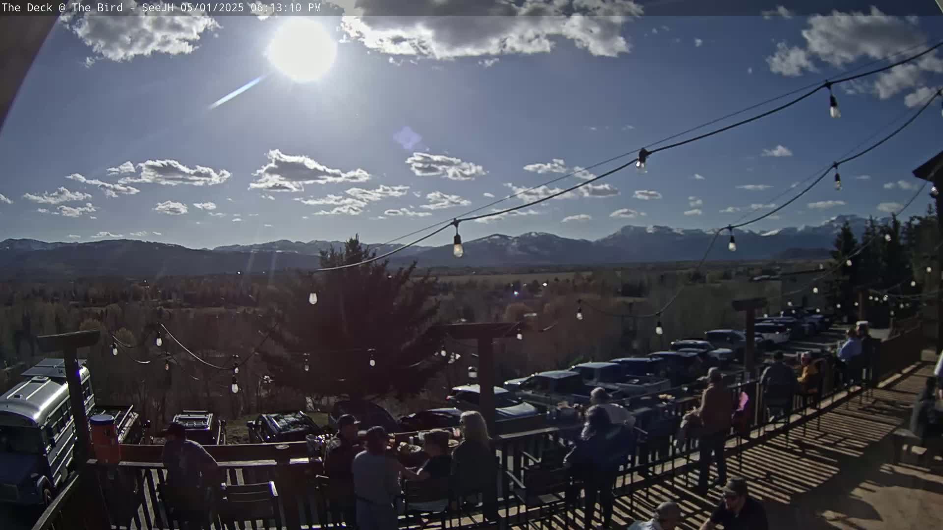 A sunny outdoor patio scene with a mountain view, string lights, and several people seated at tables.