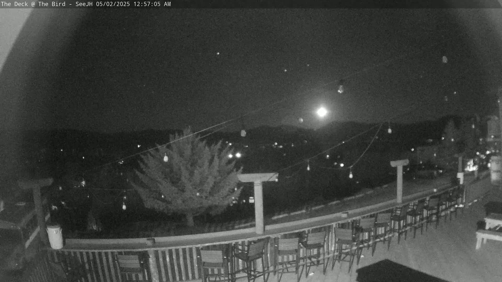 A nighttime outdoor scene shows a deck with empty chairs and a railing overlooking a tree and distant lights under a mostly clear sky.