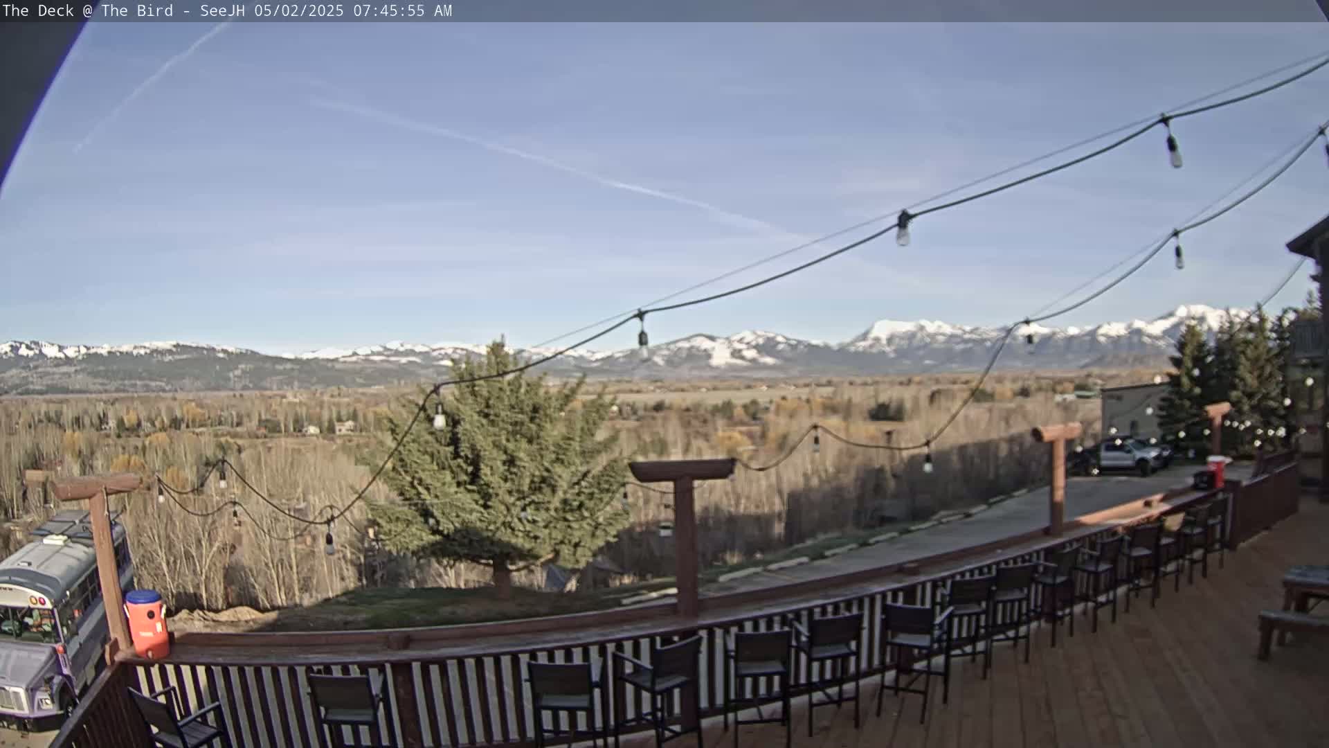 A deck with empty chairs overlooks a valley and snow-capped mountains under a clear, sunny sky.