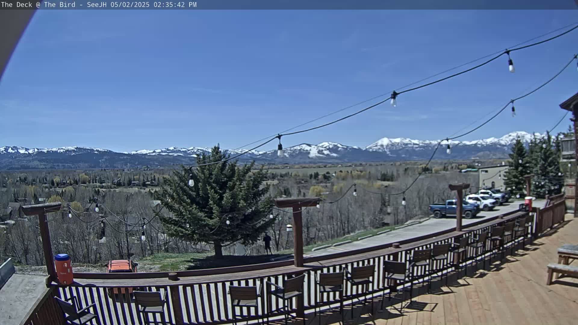 A deck with wooden chairs and a bar overlooks a valley and snow-capped mountains under a clear blue sky.
