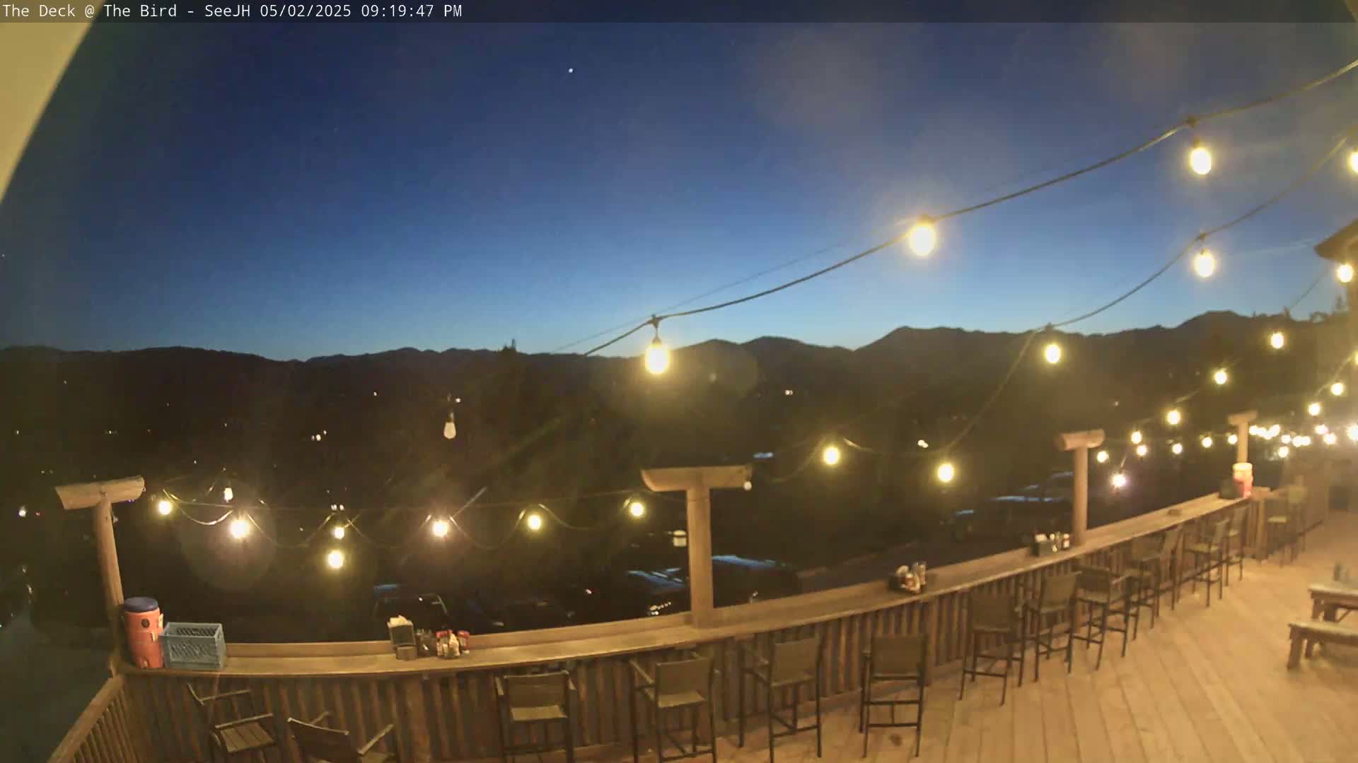 An outdoor deck at dusk, illuminated by string lights, overlooks a mountain range under a clear, dark blue sky.