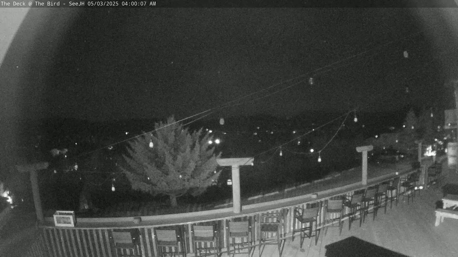 A nighttime, outdoor view shows a deck with numerous chairs and a string of lights overlooking a dark, distant town.
