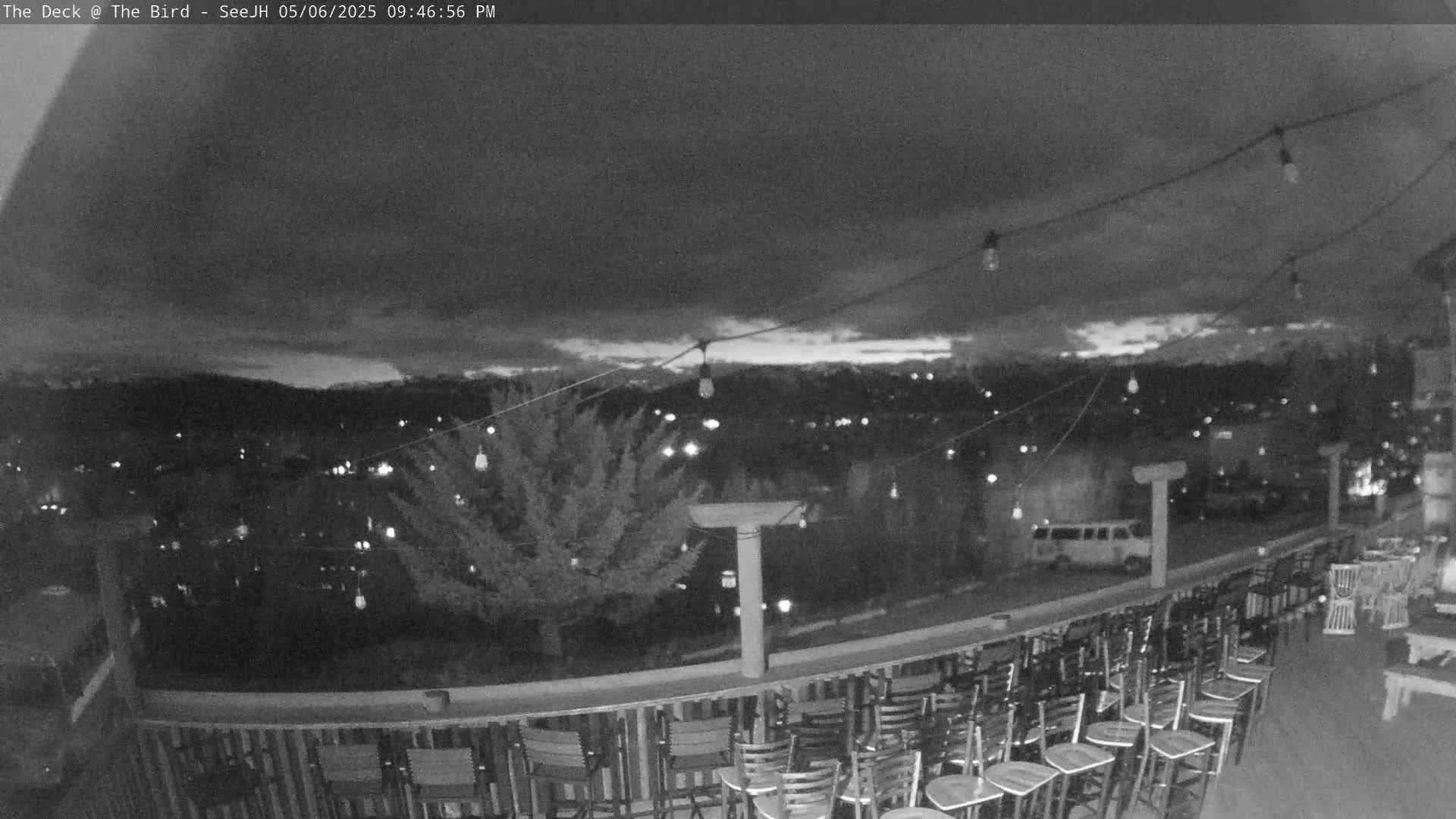 A nighttime view from an outdoor deck shows a city illuminated with lights below, string lights overhead, and numerous empty chairs lined up along a railing.