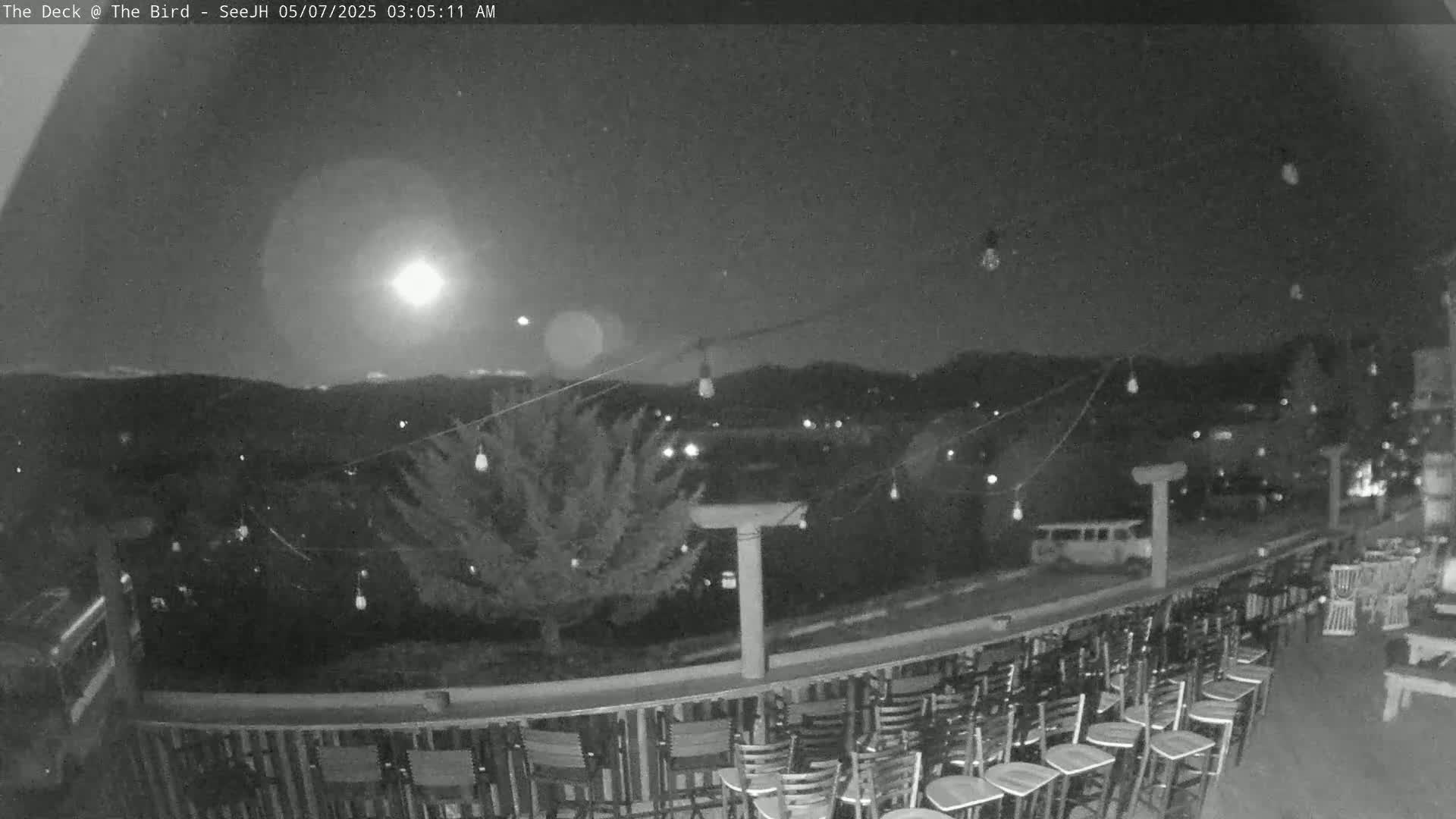 A nighttime outdoor scene shows a moonlit deck with numerous empty chairs and string lights overlooking a hilly landscape.