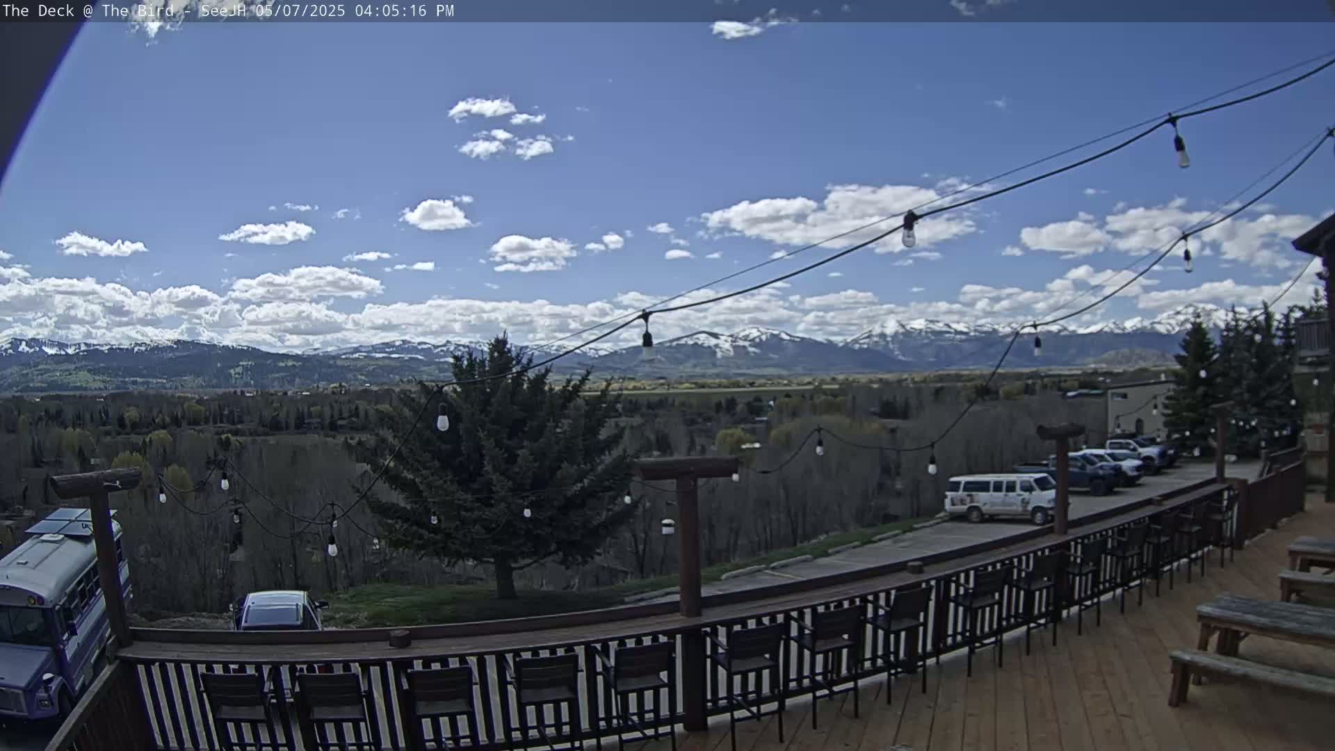 A deck with a railing and outdoor seating overlooks a valley, a few parked vehicles, and snow-capped mountains under a partly cloudy blue sky.