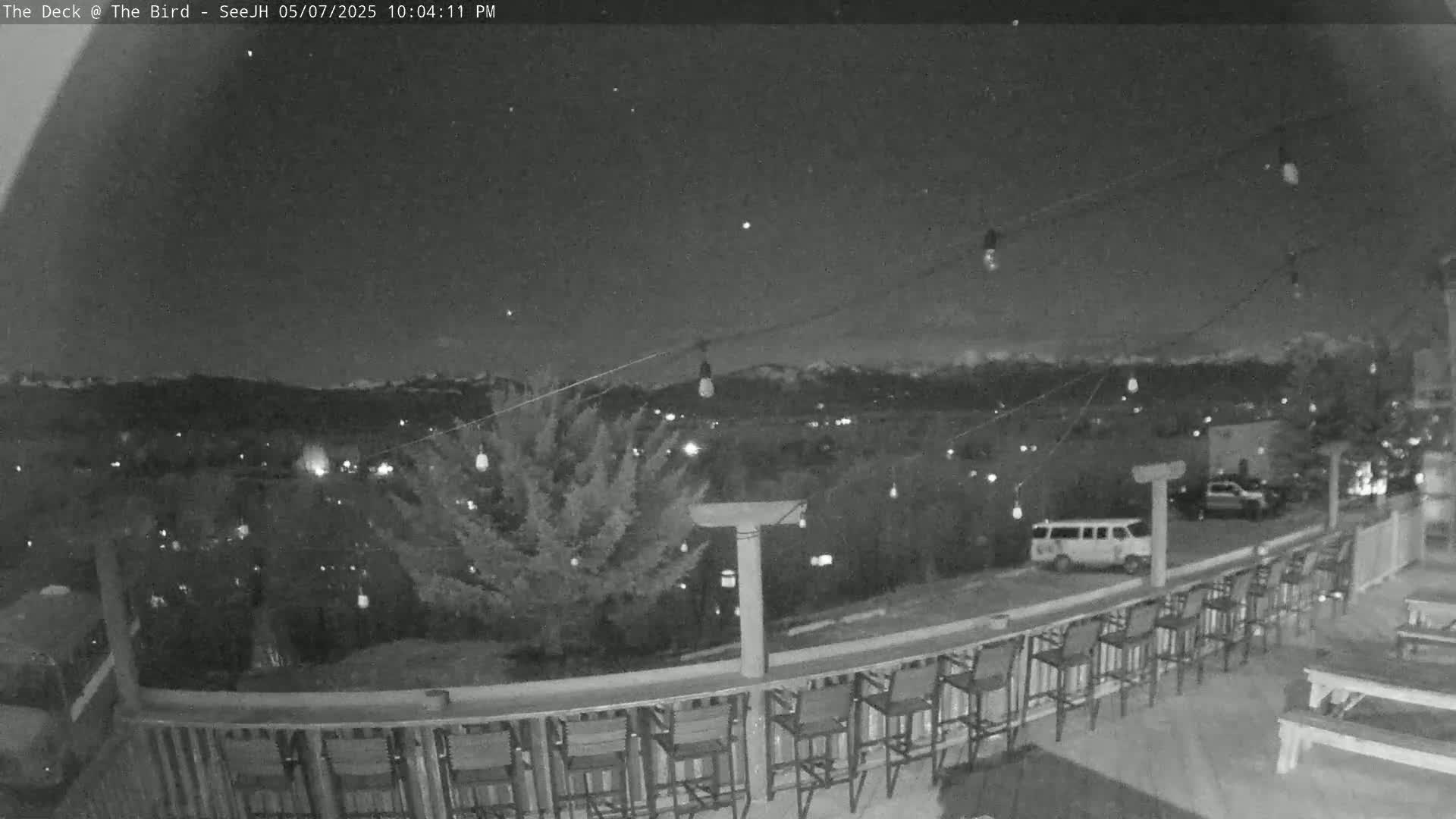 A nighttime view from a deck shows a row of empty bar stools along a railing overlooking a town and distant mountains under a clear, dark sky.