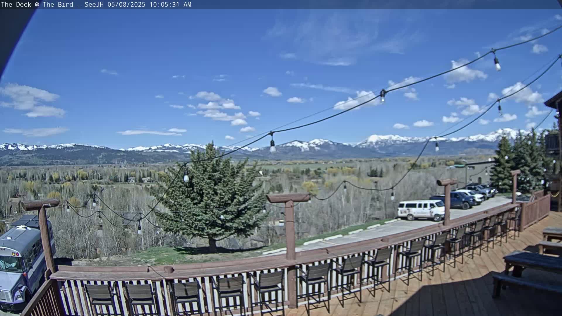 A deck with several bar stools overlooks a valley and snow-capped mountains under a partly cloudy blue sky.