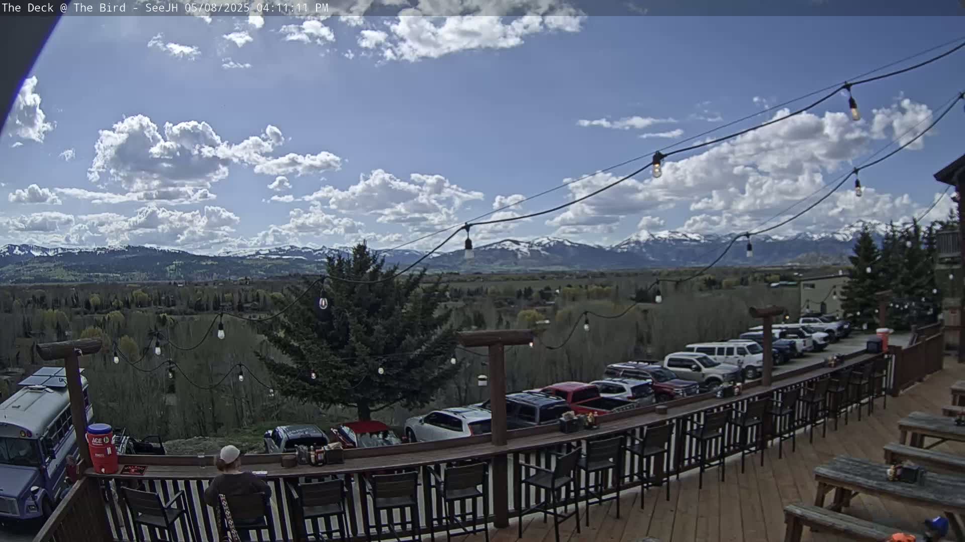 A partly cloudy day shows a mountain range in the distance, viewed from a deck with a railing and outdoor seating, cars parked below, and string lights overhead.