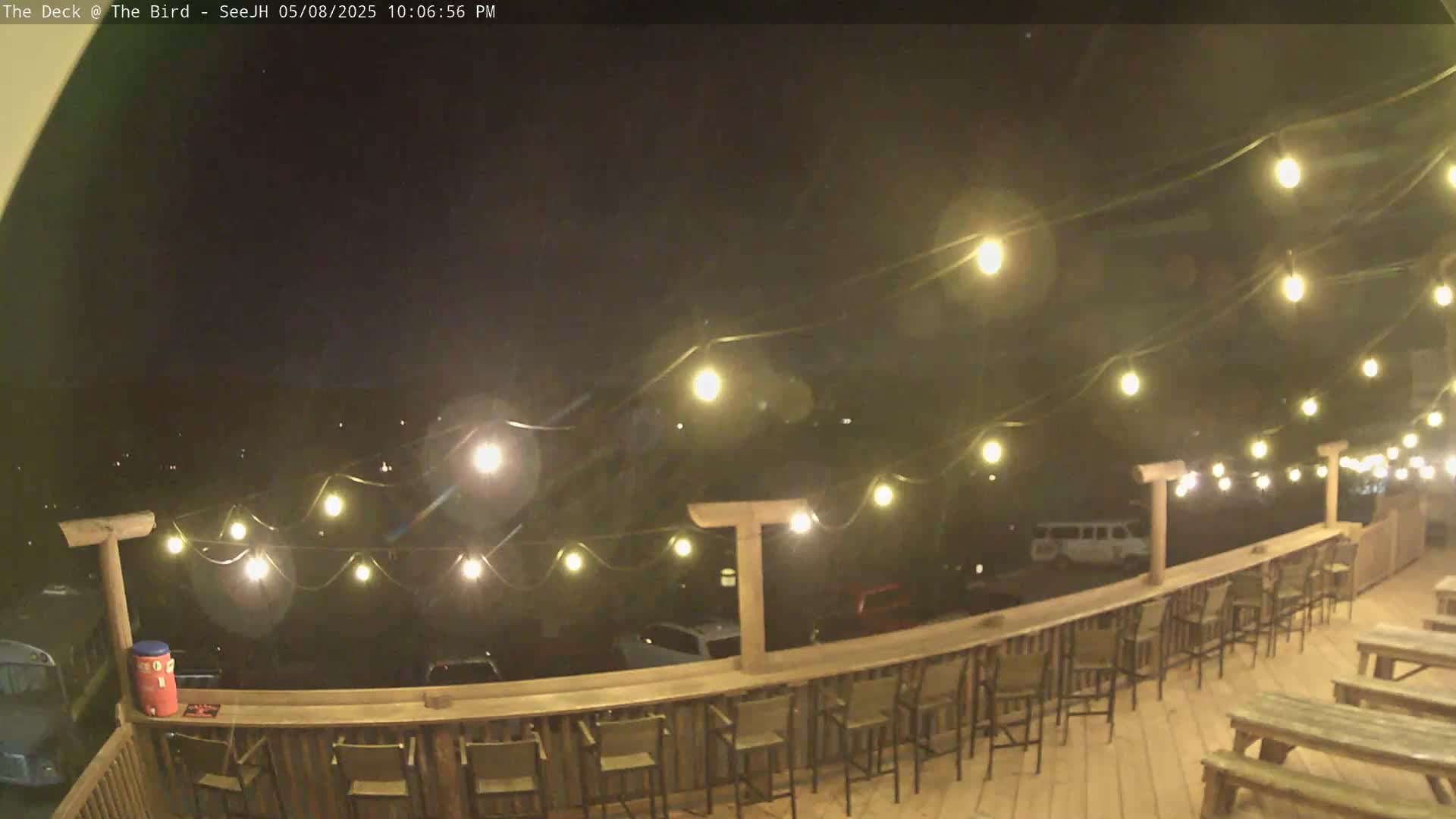 A nighttime outdoor deck with string lights overhead, wooden tables and chairs, and a railing overlooking a parking lot.